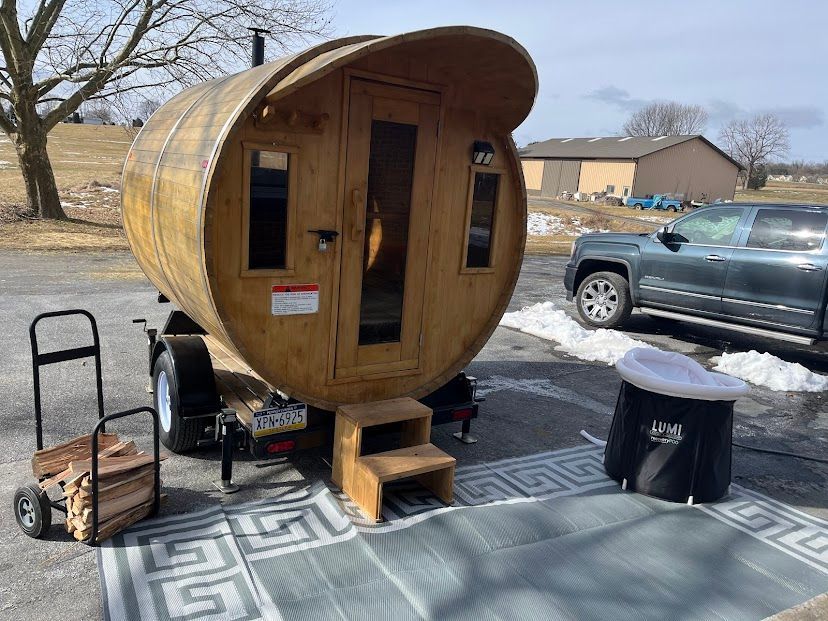 Small wooden pod cabin on a trailer at an outdoor lot, with chairs, a table, and a parked pickup nearby.