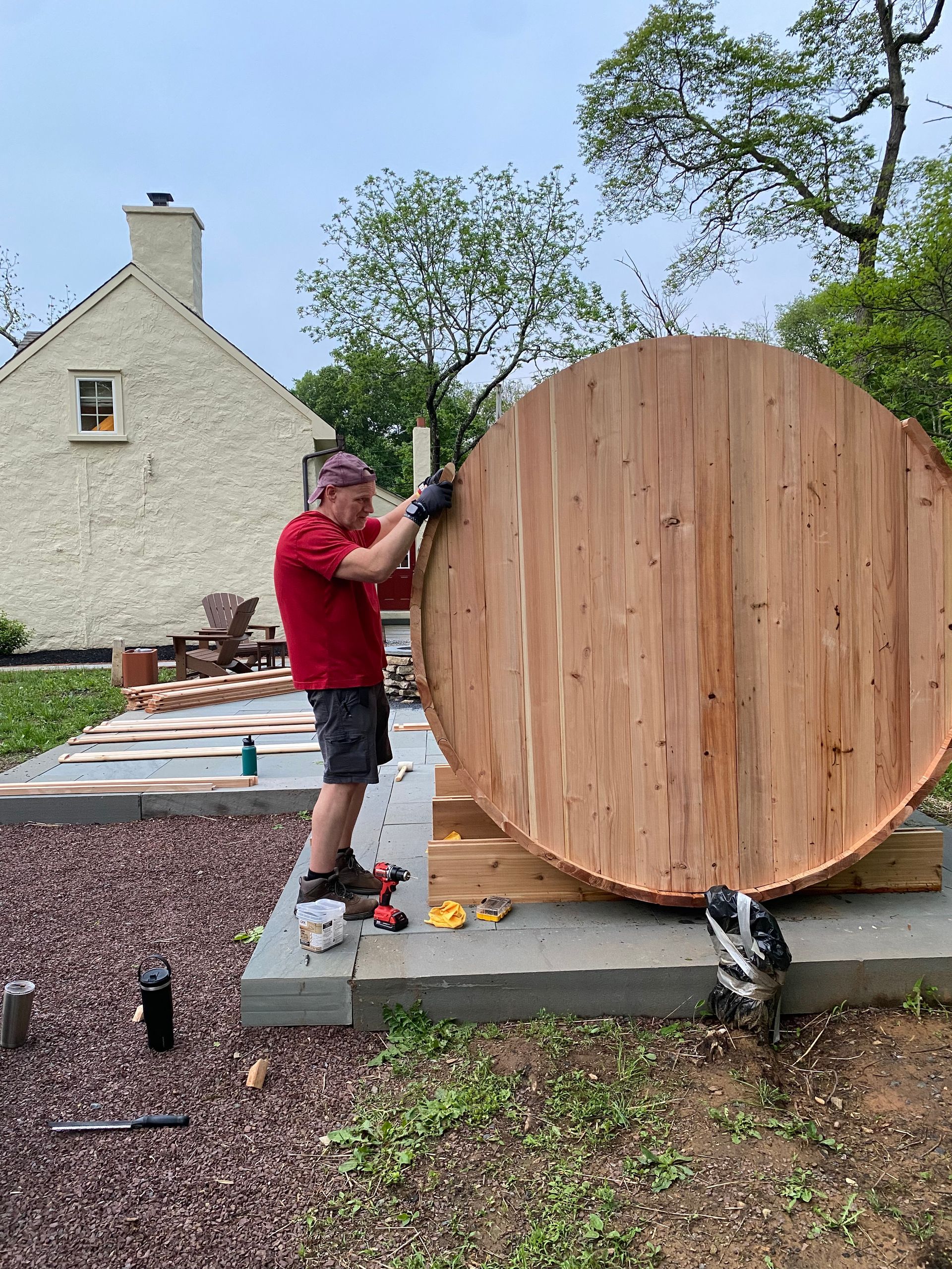Man in red shirt standing beside a large wooden round structure outdoors beside a stone house