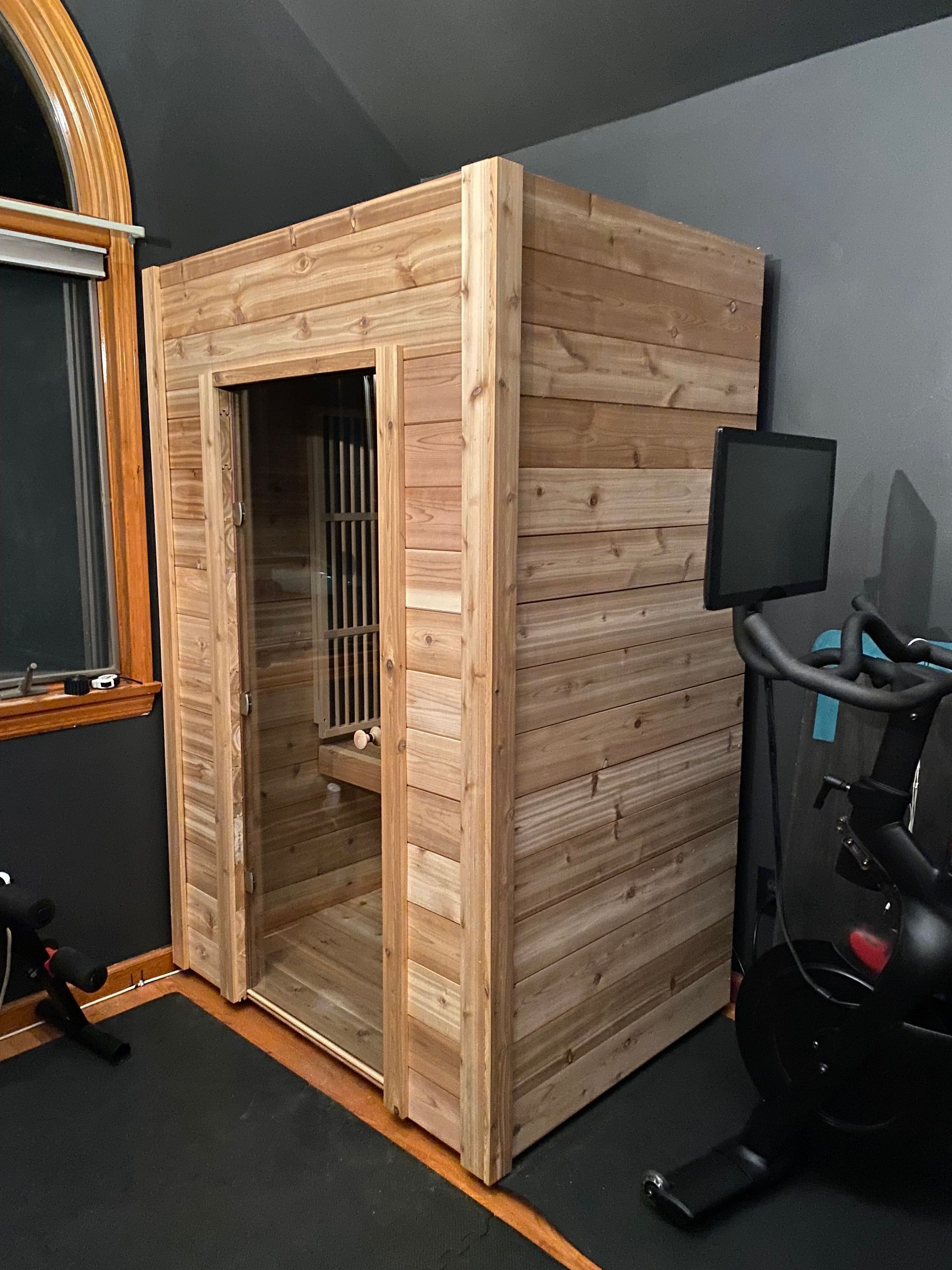 Wooden sauna room in a gym with an open door, beside exercise equipment on dark flooring.