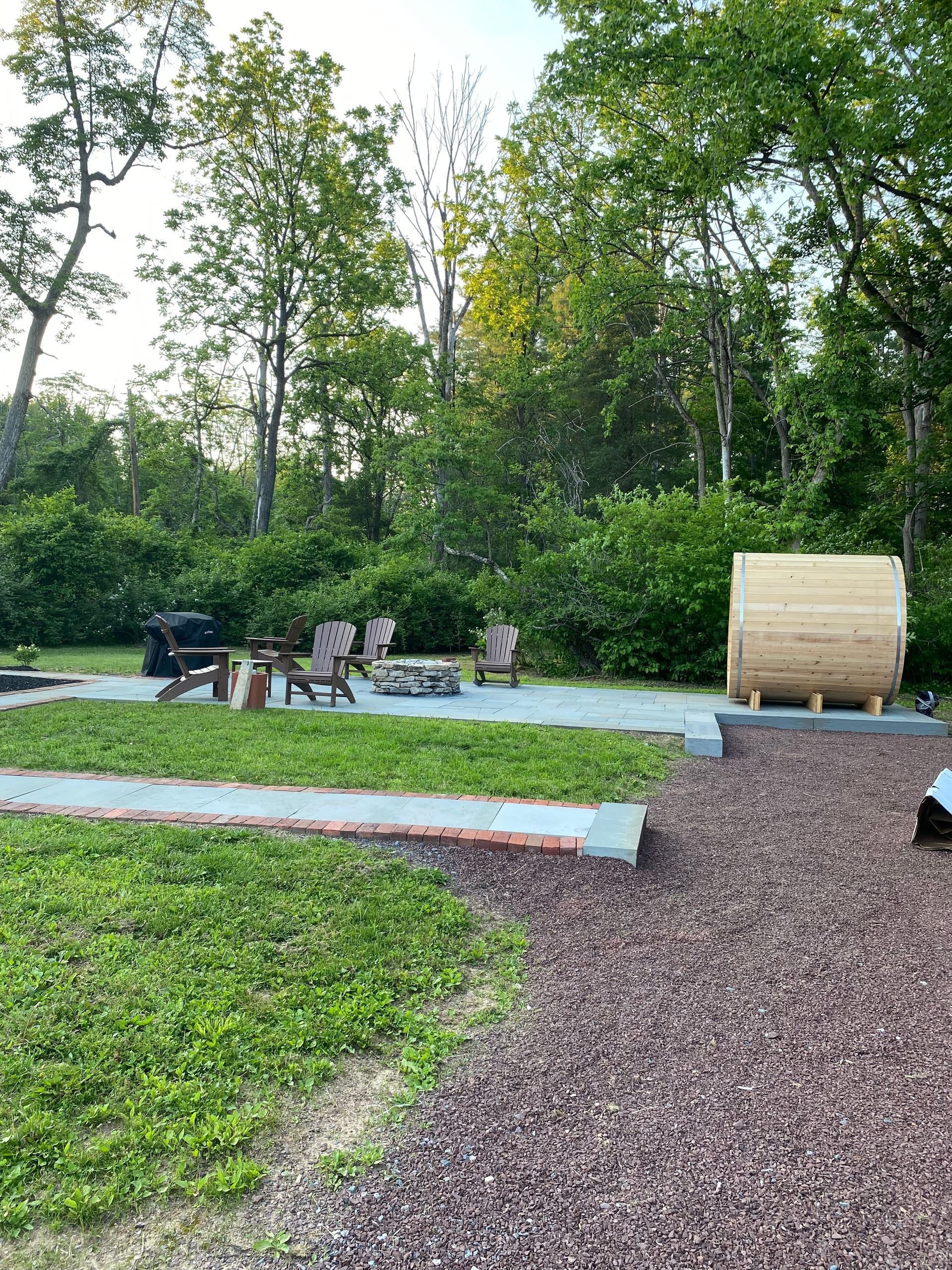 Park with a fire pit, benches, and a large wooden spool on a gravel path surrounded by trees.