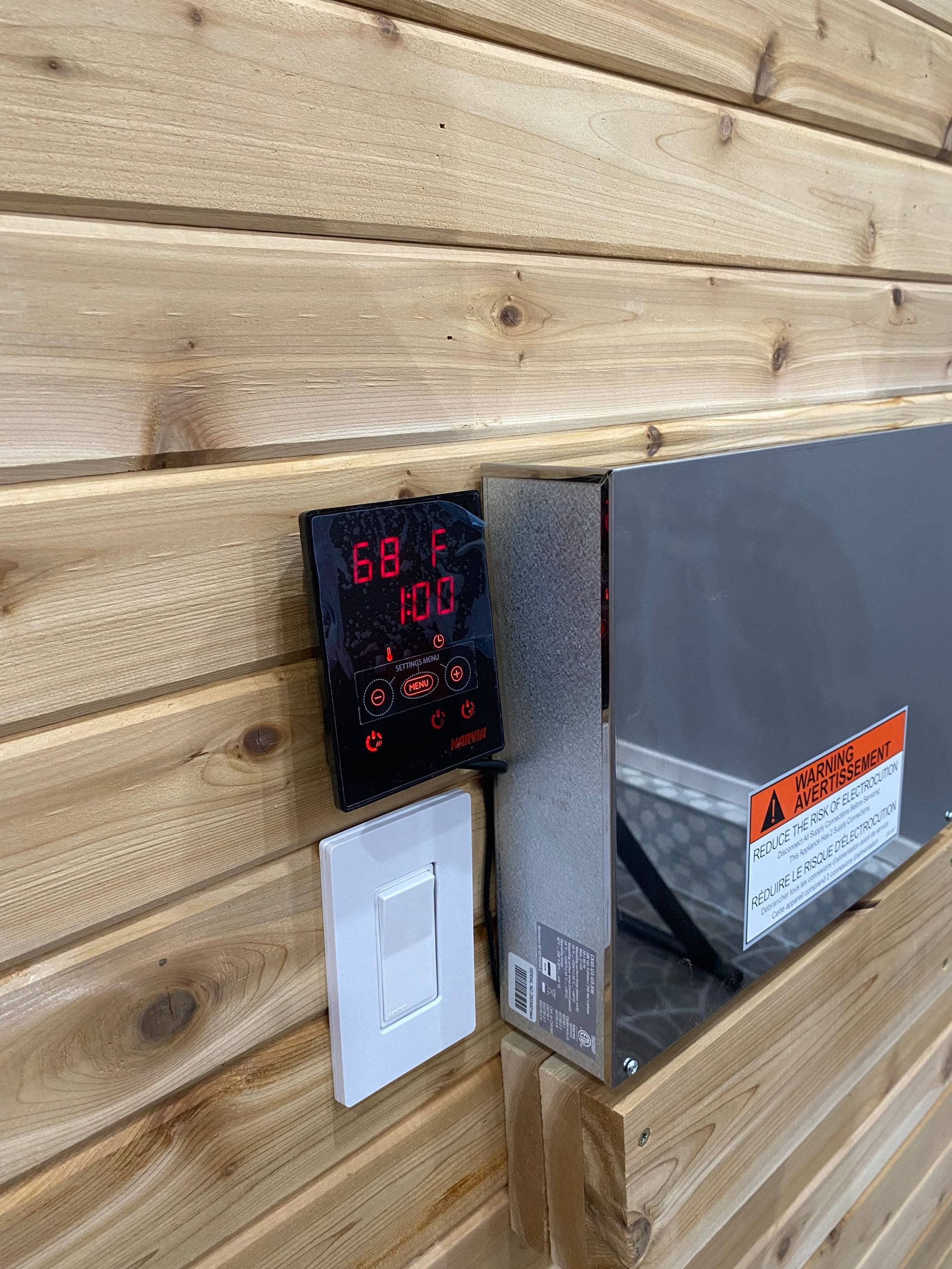 Wall-mounted digital display with red numbers, next to a light switch and a gray metal box on wood siding.