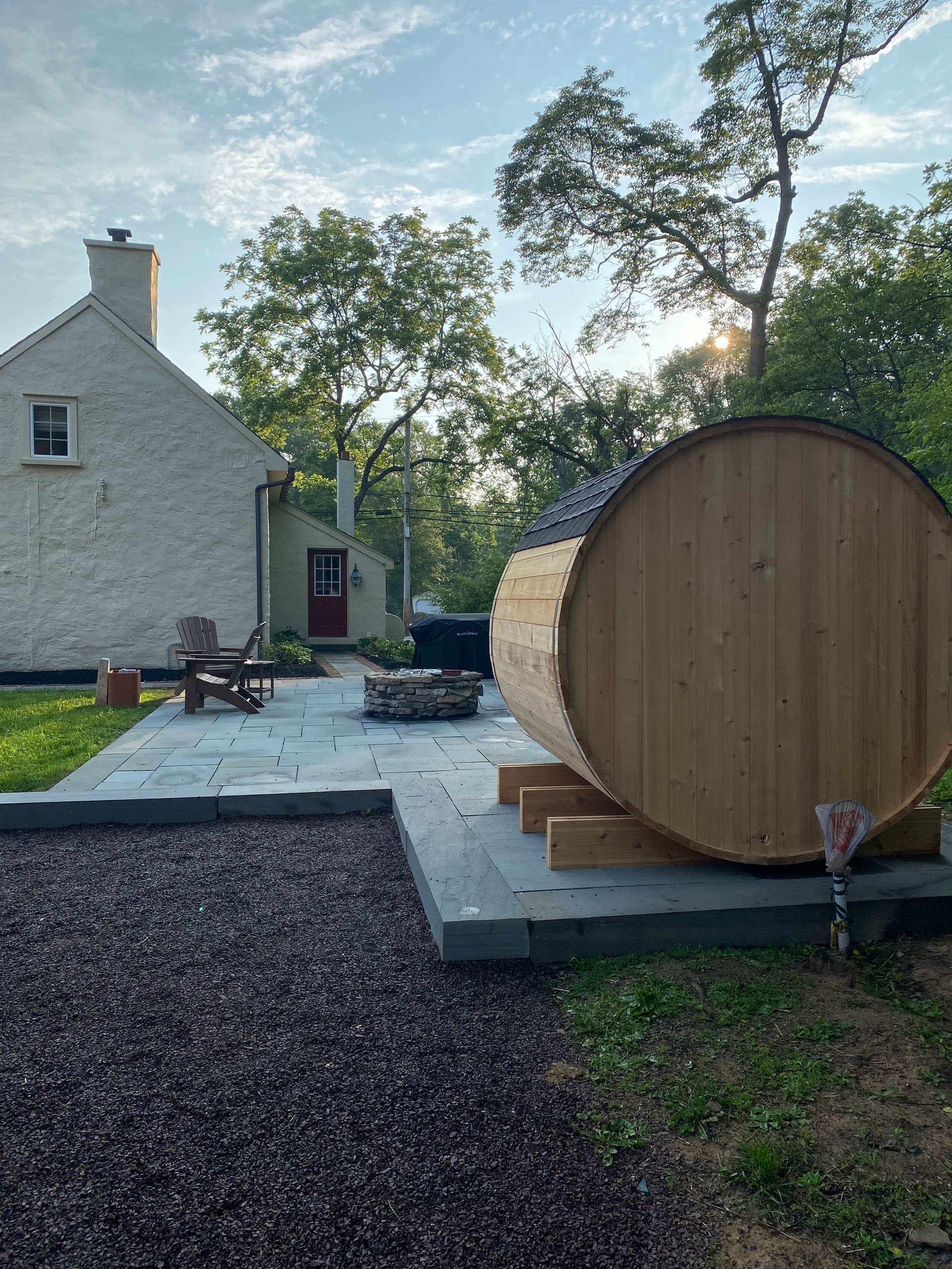 Backyard with stone patio, house, trees, and a large wooden barrel sauna on a platform