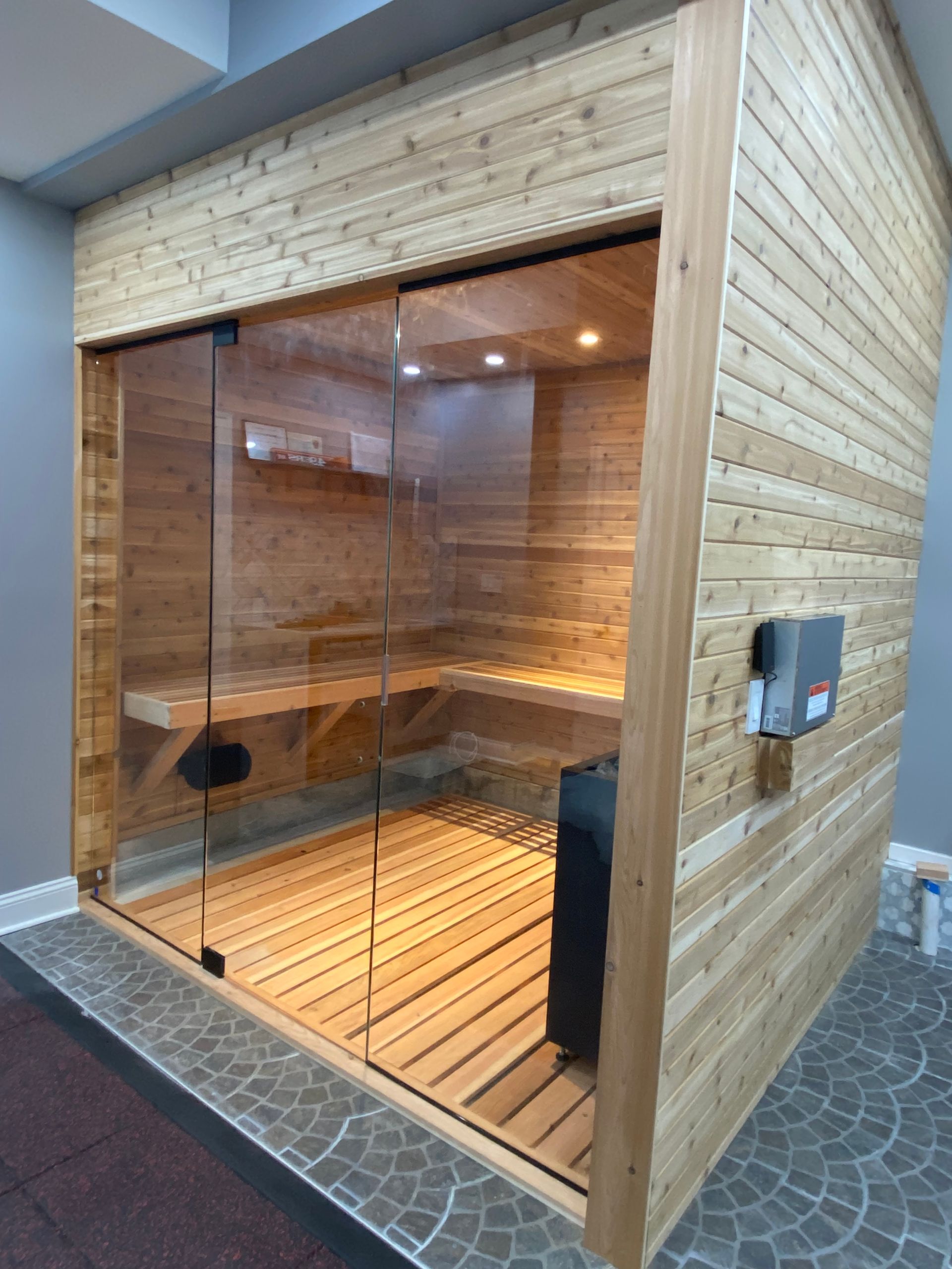 Wood-paneled glass sauna room with slatted wooden floor and interior bench lighting.