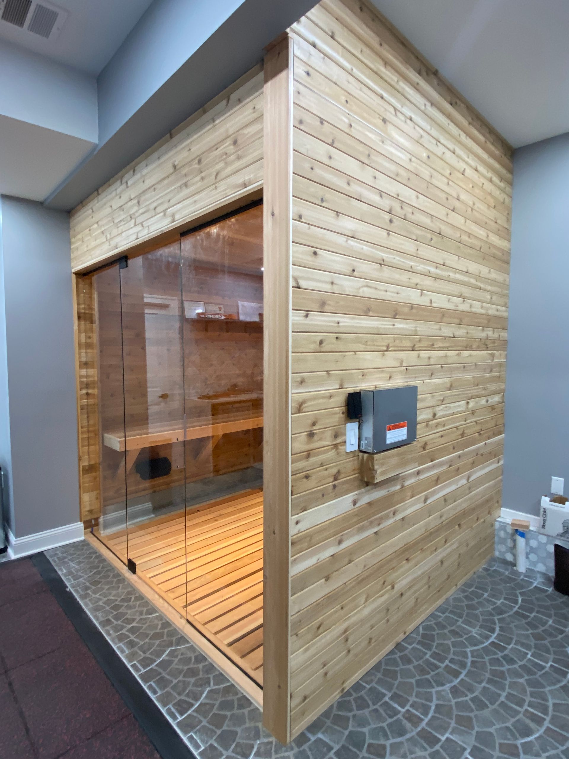 Wood-paneled sauna room with a glass door and exterior control panel, set in a tiled indoor area