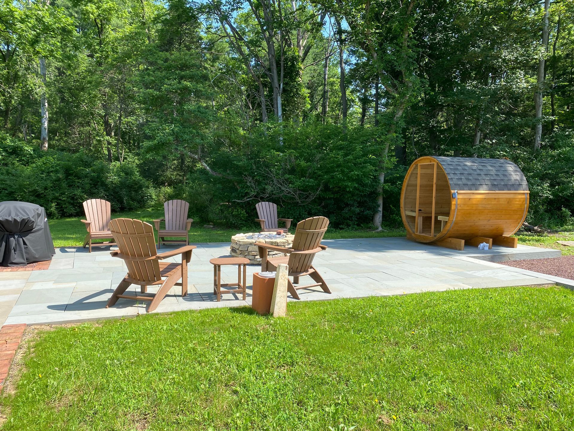 Outdoor patio with wooden chairs, fire pit, and a barrel sauna beside a wooded lawn.