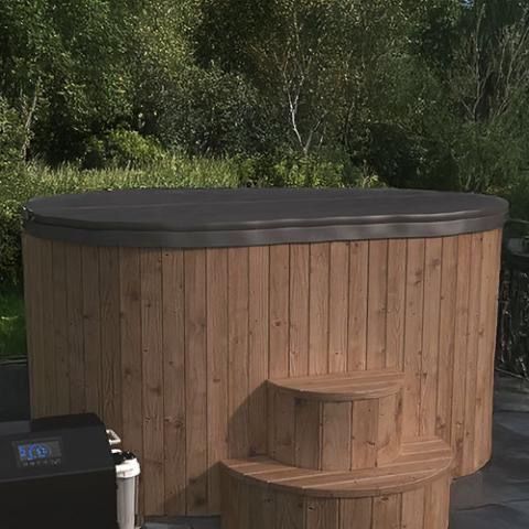Oval wooden 2 person cold plunge tub with black cover and built-in steps outdoors beside greenery.
