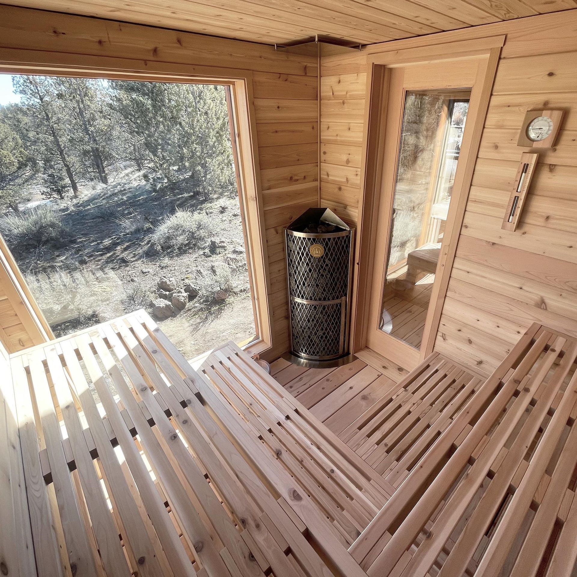 Wooden sauna interior with slatted benches, a heater, and a window looking onto snowy trees