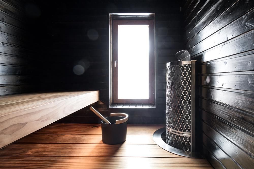 Sauna interior with wooden bench, bucket, and heater beside a bright window