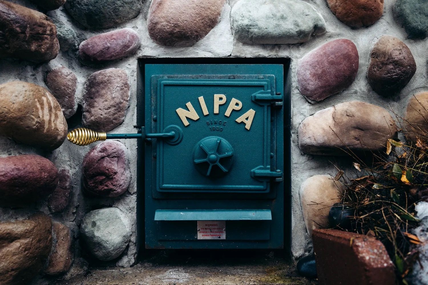 Blue metal NIPA door set in a stone wall with a brass handle and surrounding rocks and plants