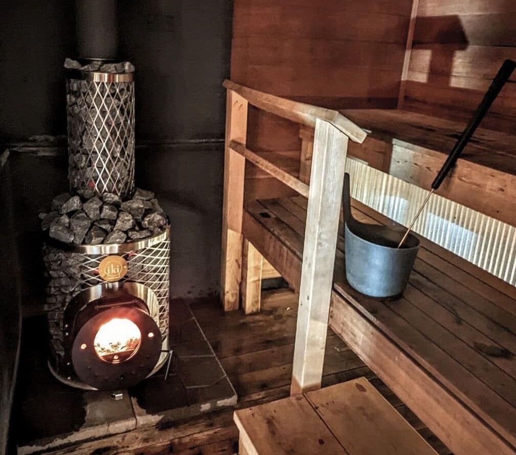 Rustic wood sauna with a lit metal stove and a bucket beside a bench