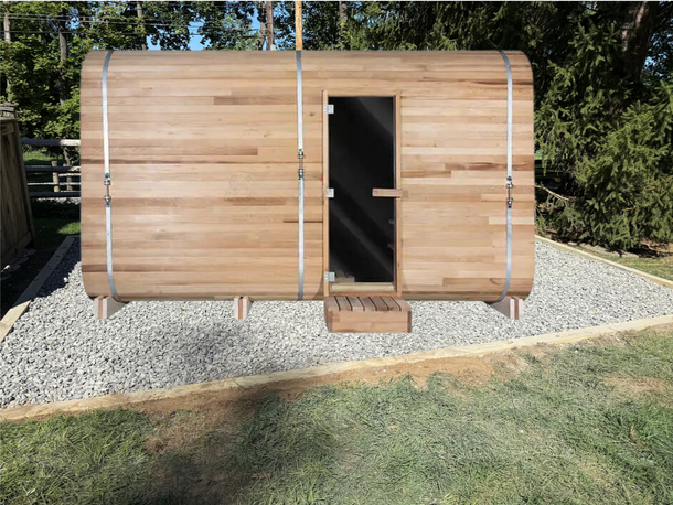 Small wooden outdoor cabin with a door, set on gravel with grass and trees around it.