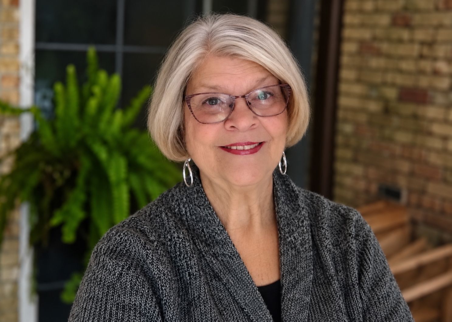 Portrait of a smiling woman with glasses, gray hair, and a patterned top in front of leafy greenery