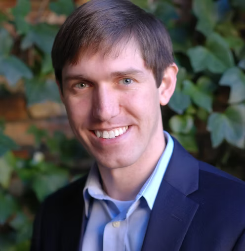 Smiling man in a navy blazer and blue shirt against a leafy background