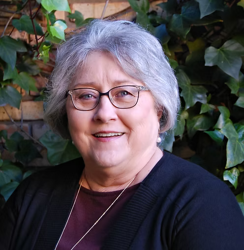 Smiling person with glasses in a black cardigan, standing in front of leafy greenery.