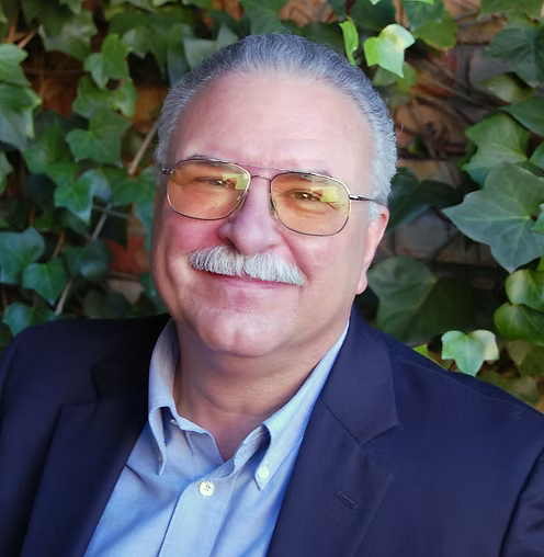 Portrait of a smiling man with glasses and a mustache in a navy blazer, standing before leafy greenery