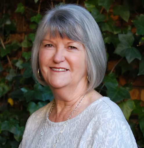 Smiling woman with gray hair wearing a white lace top against a leafy green background