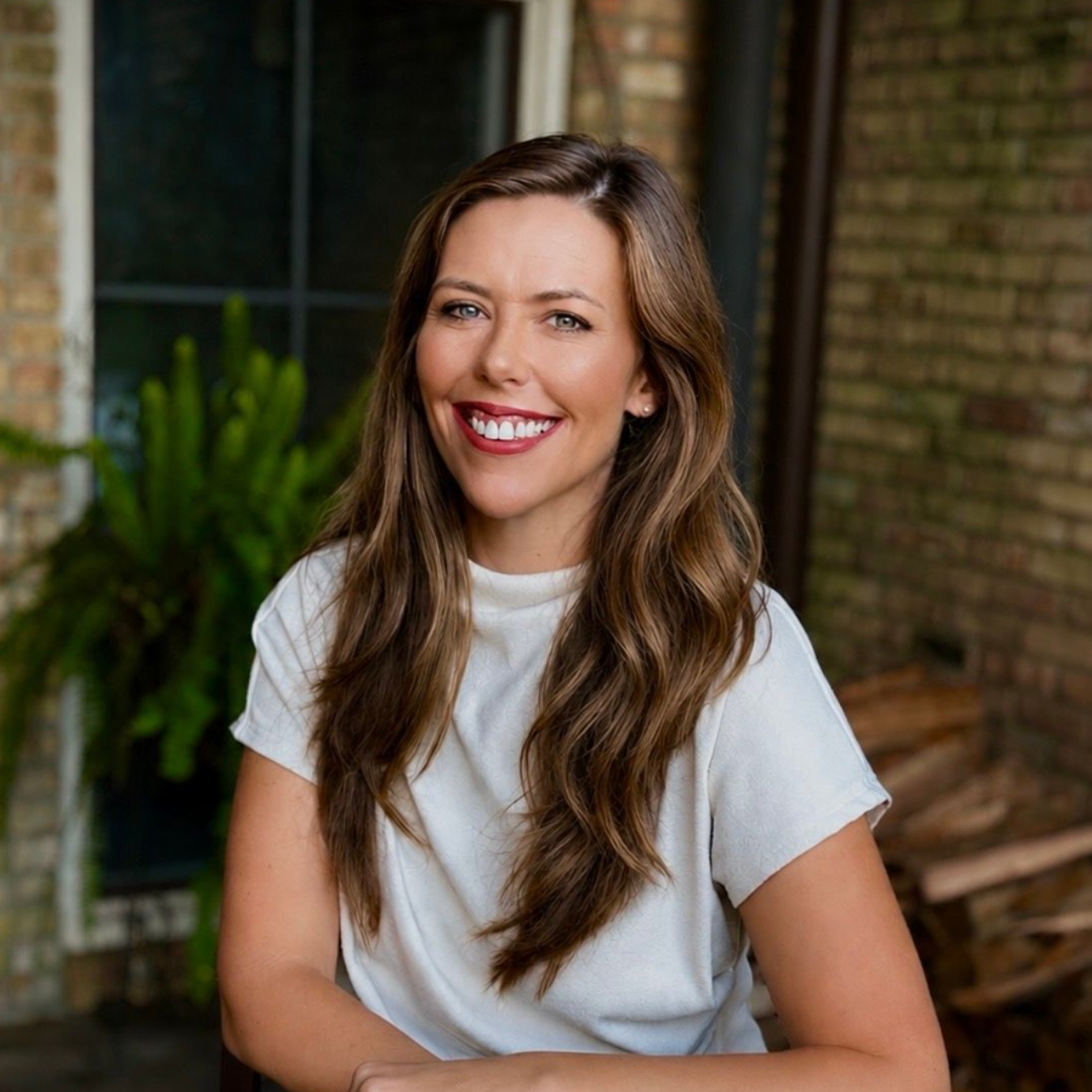 Smiling woman in a white shirt seated outdoors by a brick wall and potted plant