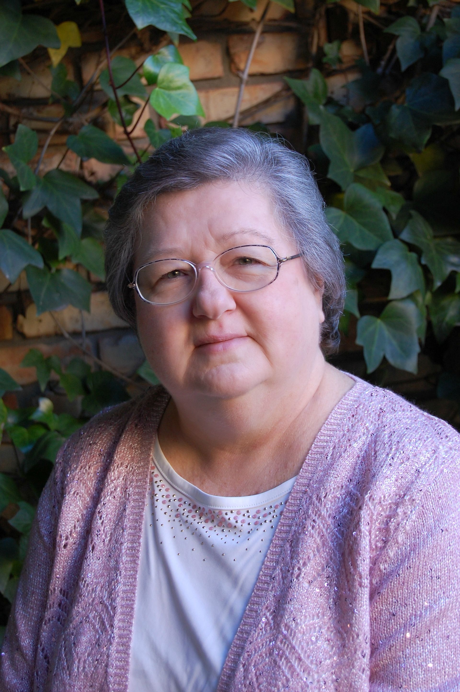 Portrait of a woman with glasses and short gray hair, wearing a lavender cardigan in front of ivy-covered brick.