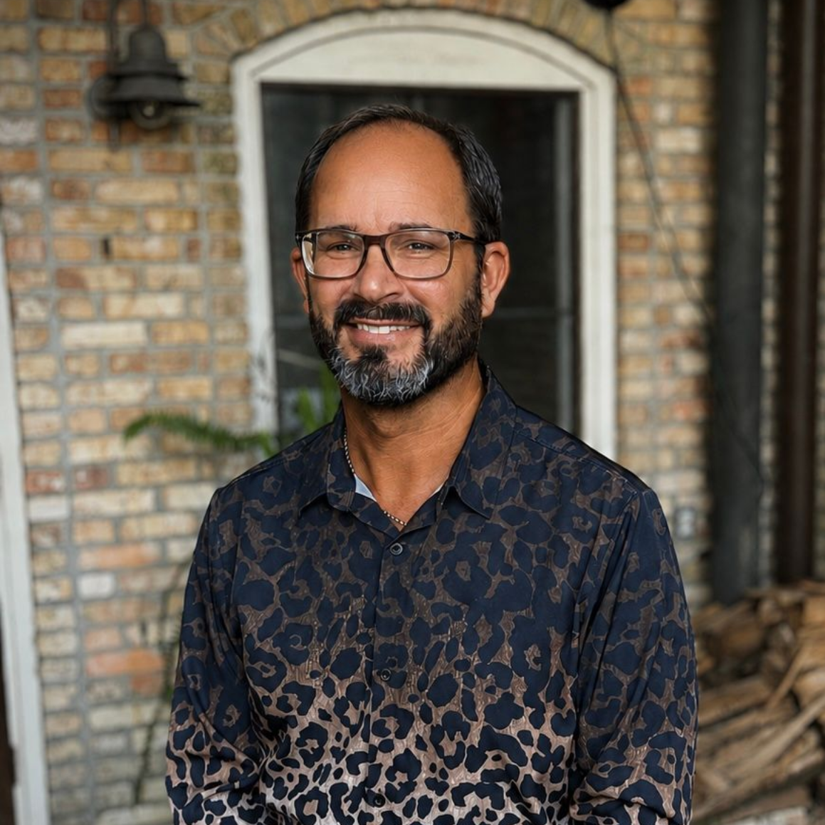 Man smiling in a patterned dark shirt, standing in front of a brick house doorway.