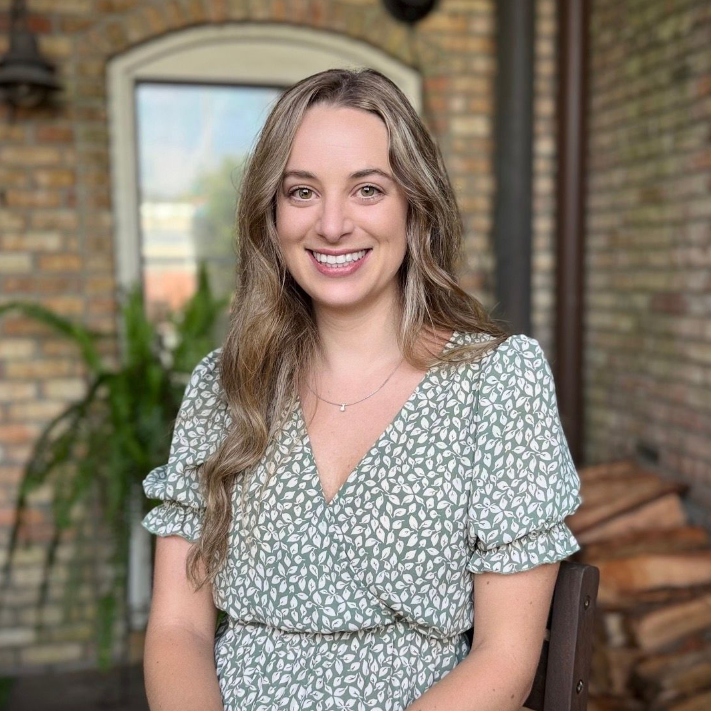 Smiling woman in a green floral dress seated on a porch with brick walls and an arched window behind her