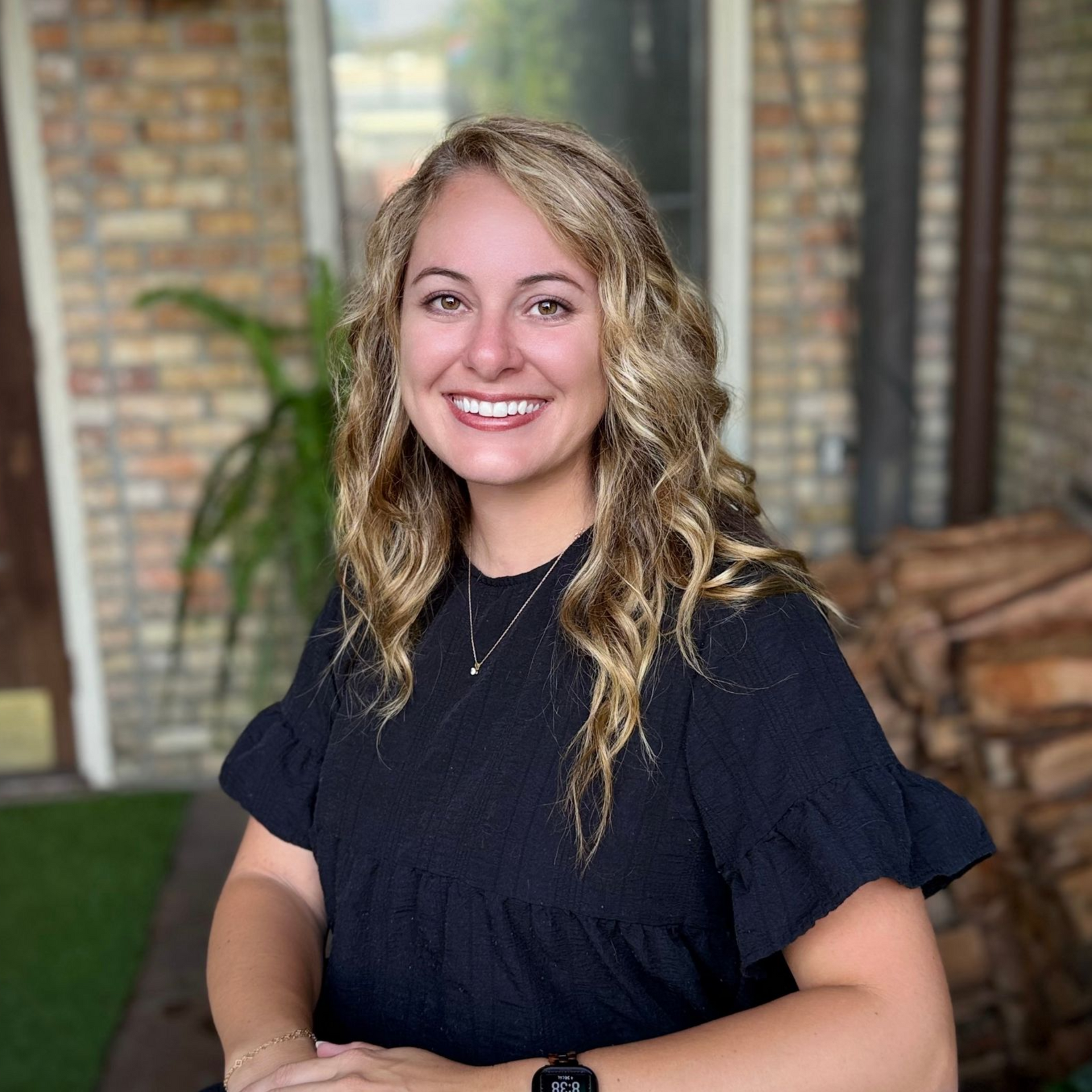 Smiling woman in a dark top standing outdoors in front of a brick wall and greenery