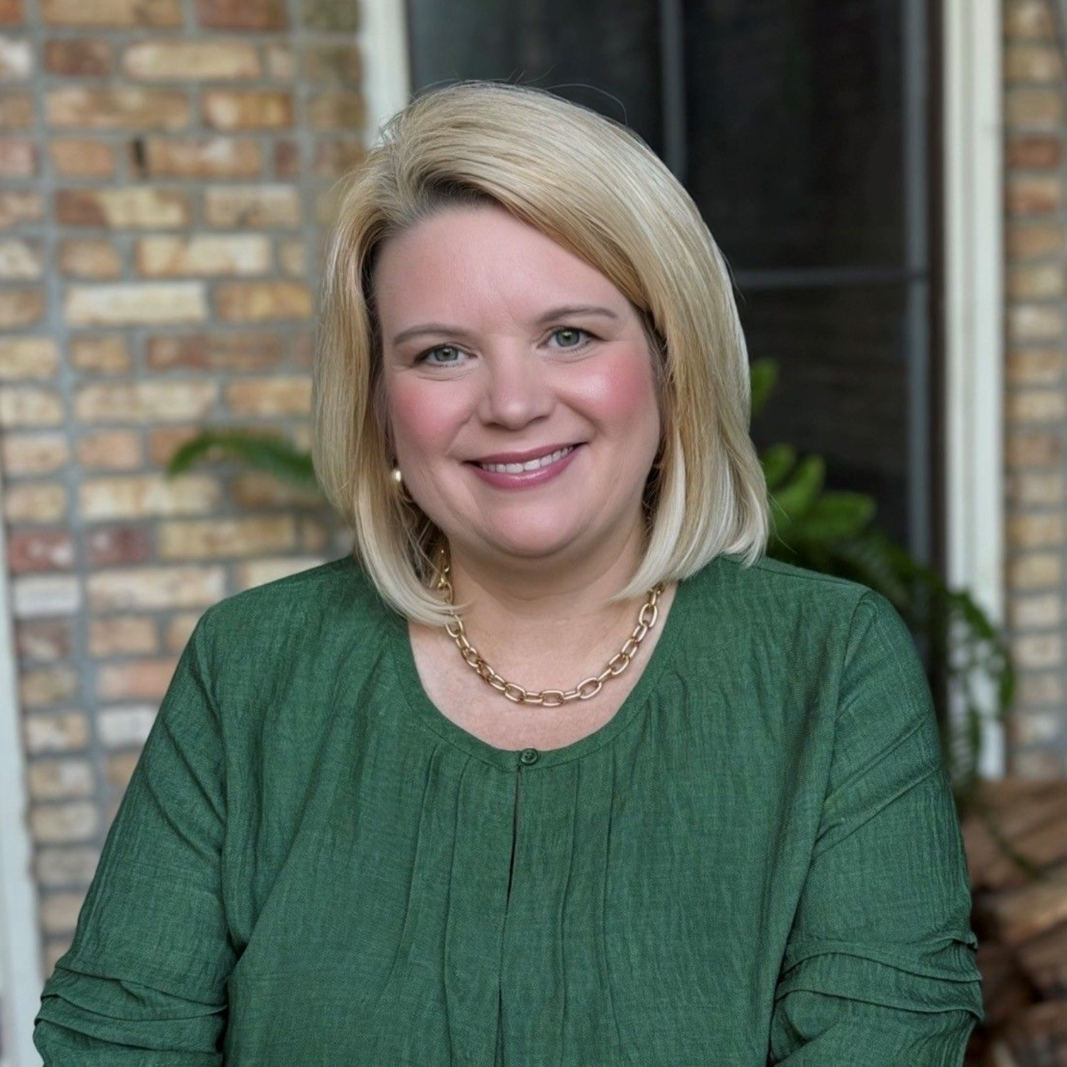 Smiling person with blonde hair in a green top, standing outdoors by a brick wall and window.