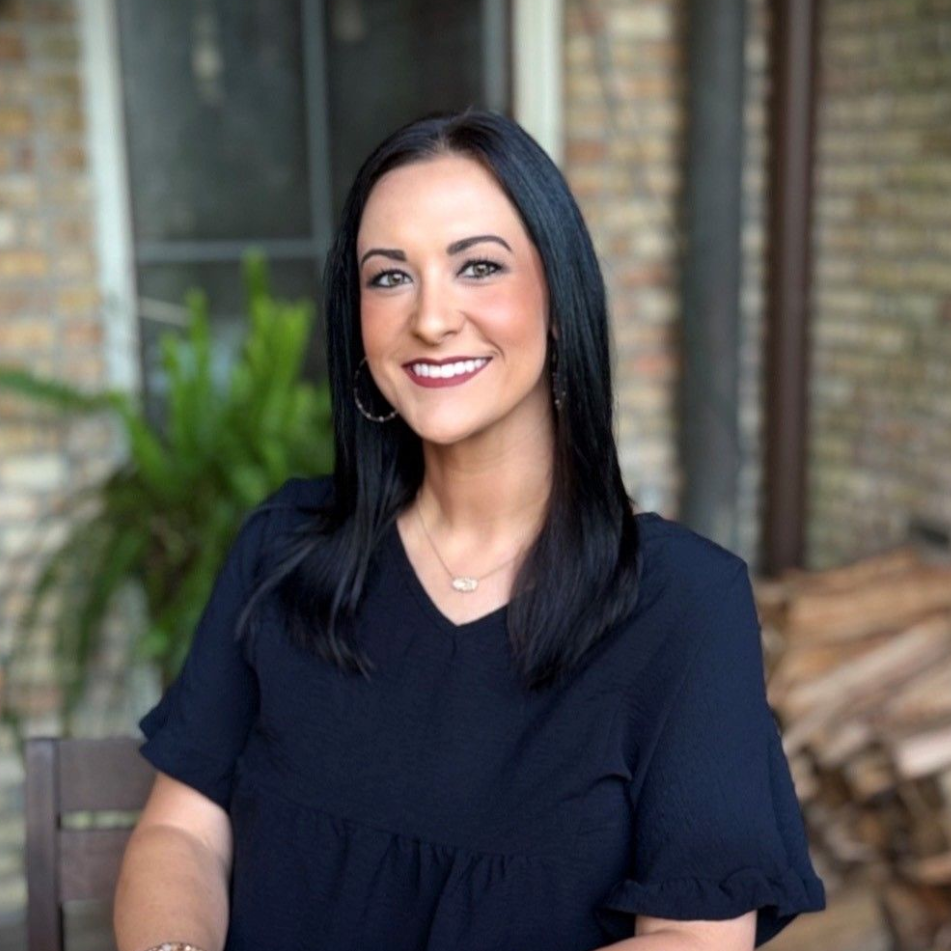 Smiling woman with straight black hair, wearing a black top, standing outdoors by a brick wall and plants