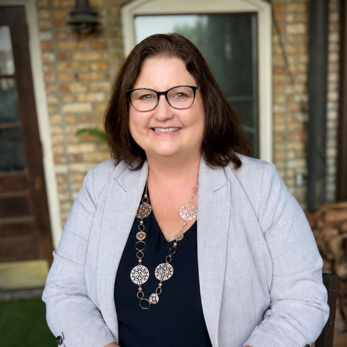 Smiling woman in glasses and a gray blazer standing outdoors in front of a brick building