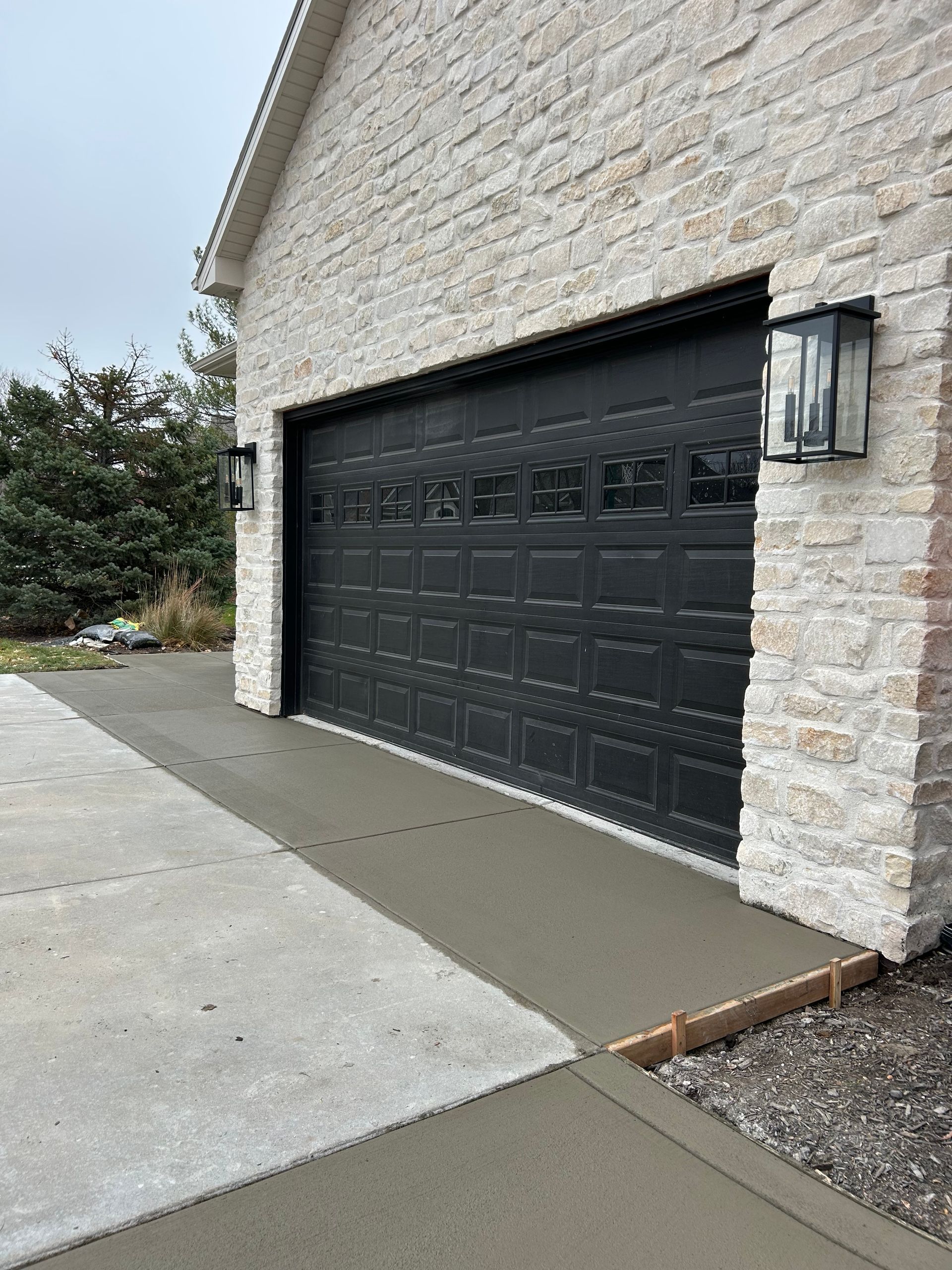 Stone house exterior with black garage door and concrete driveway, viewed from the side