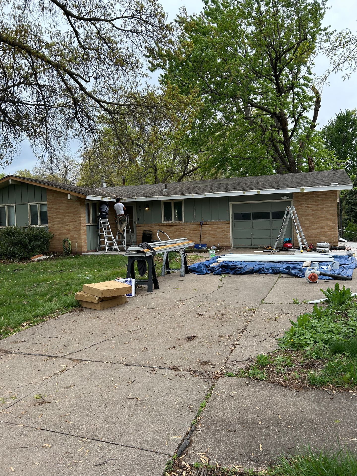 Front yard with a driveway, piled blue tarps, and scattered boxes outside a house under trees