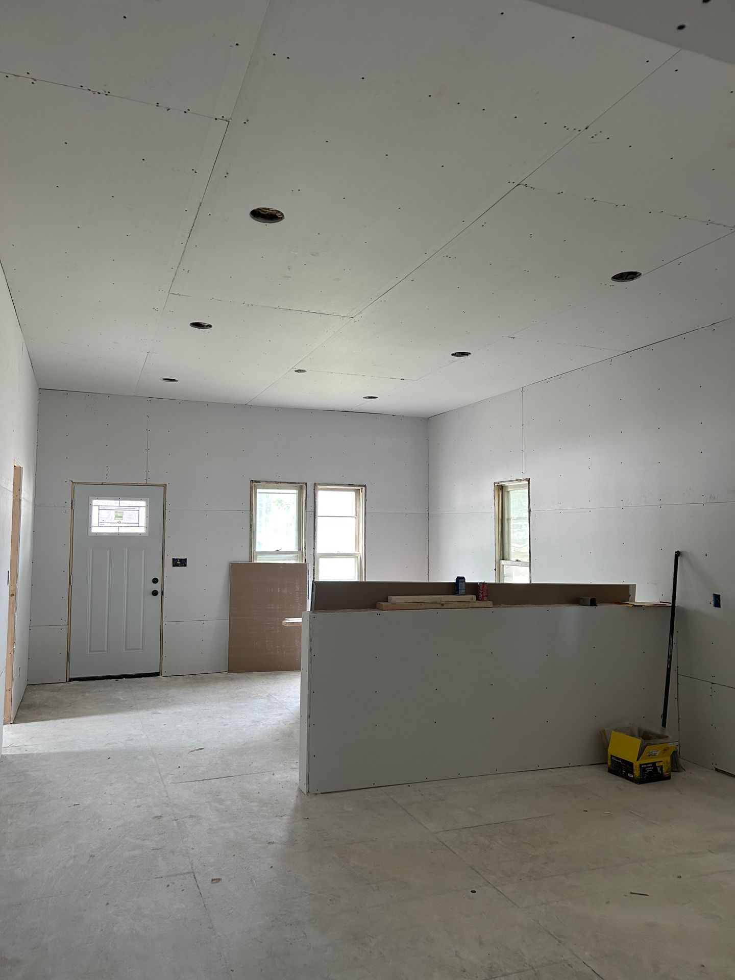 Empty unfinished room with white drywall, exposed ceiling lights, a door, windows, and a half-built partition wall.