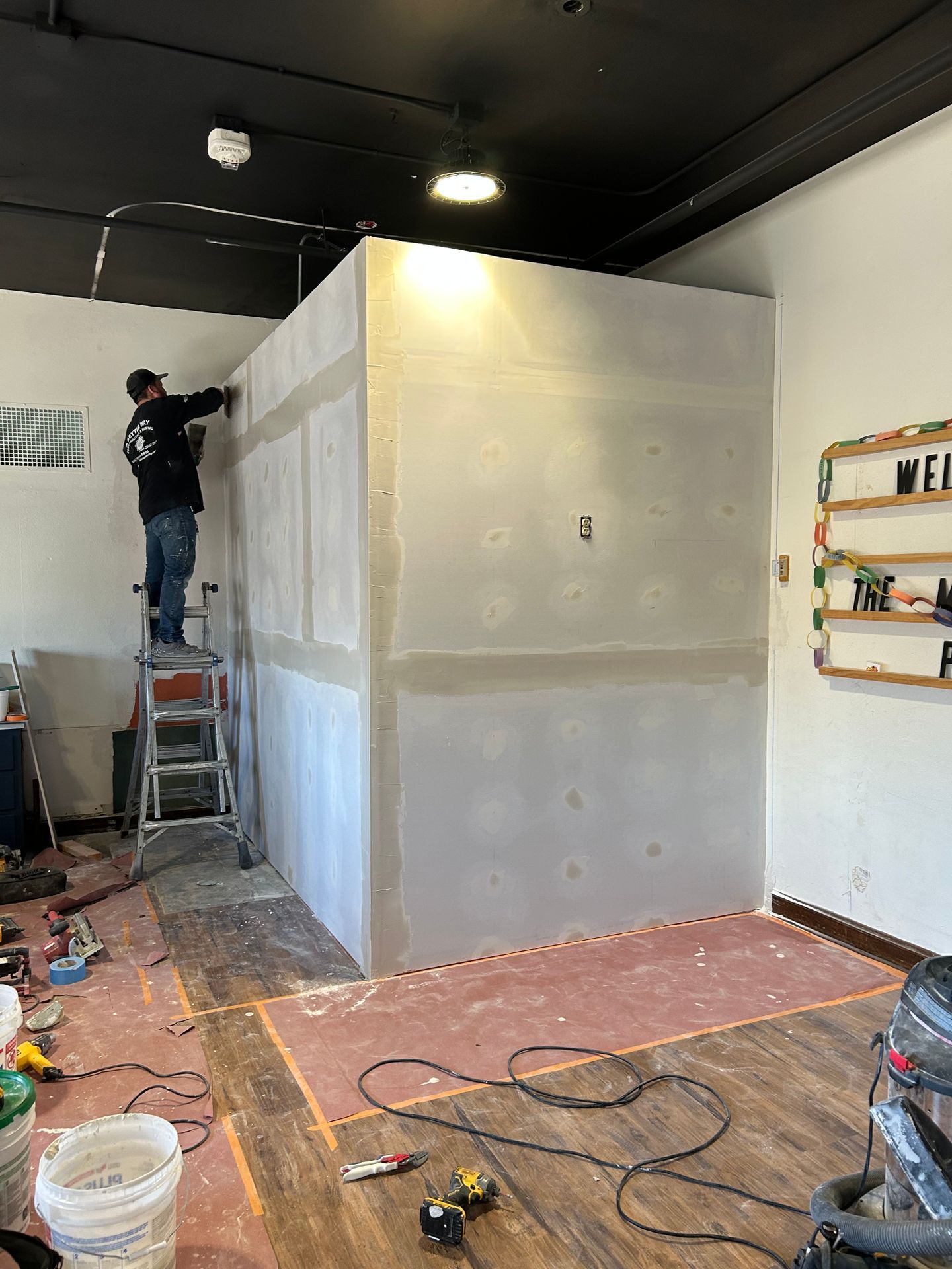 Worker on a ladder painting a large white room partition inside an unfinished interior.