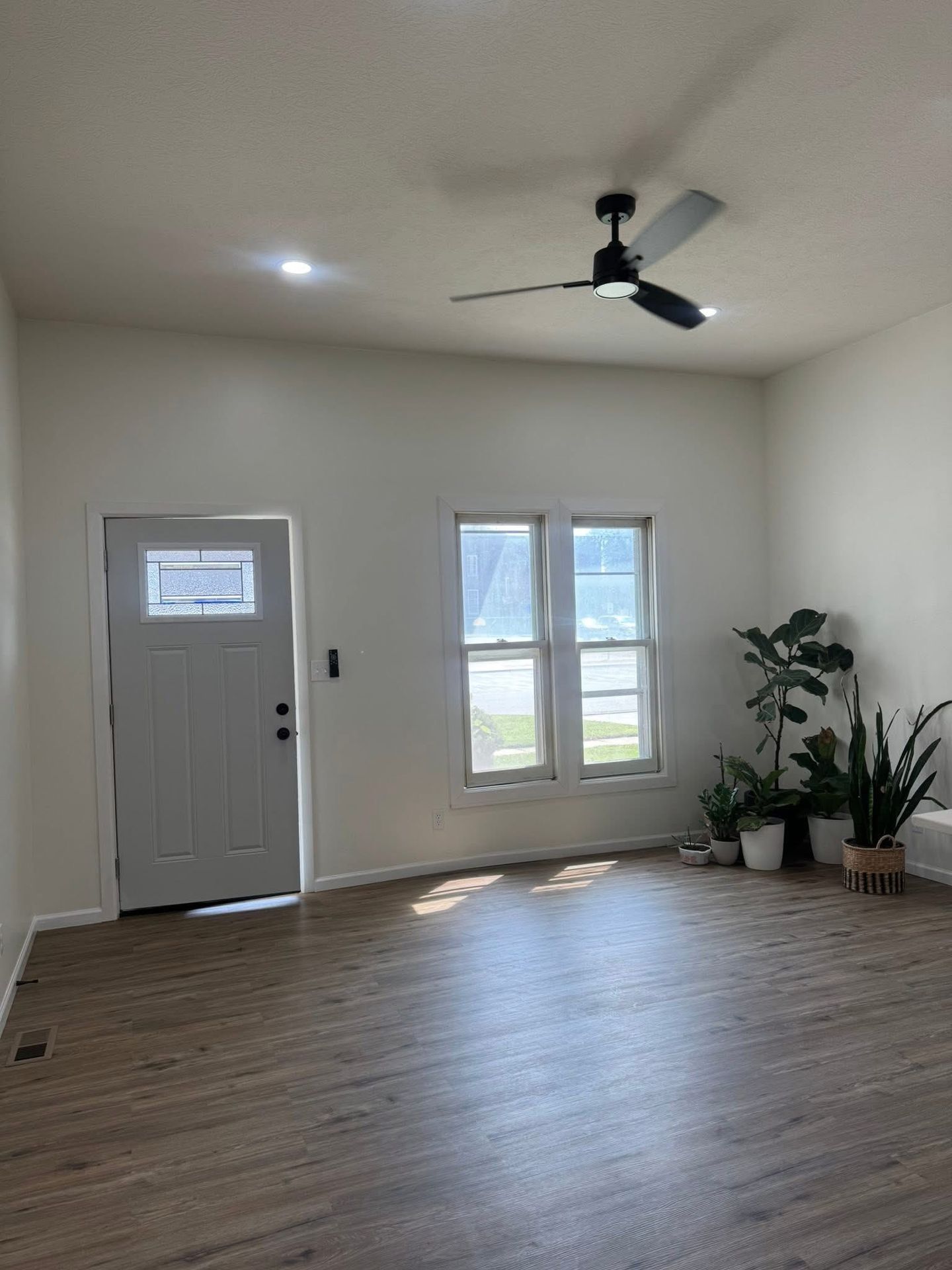 Empty living room with white walls, wood-look floor, front door, windows, ceiling fan, and potted plants.