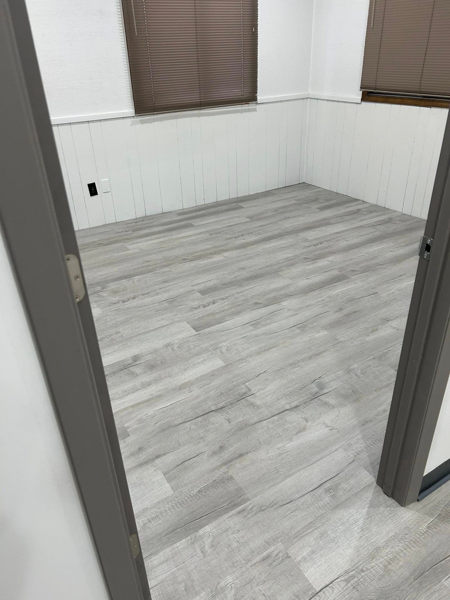 Empty room with light gray wood flooring, white paneled walls, and two curtained windows