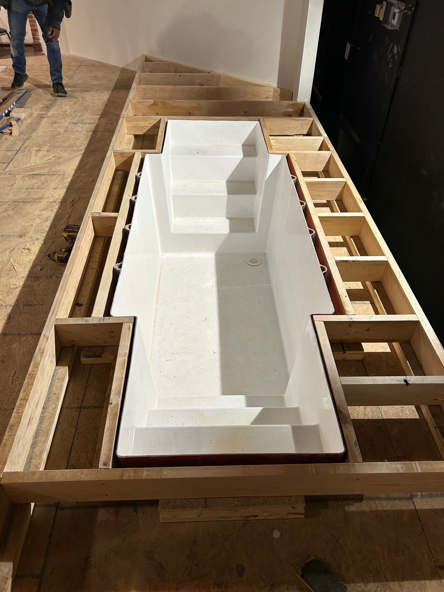 Wooden frame with a large white bathtub or shower pan being installed indoors