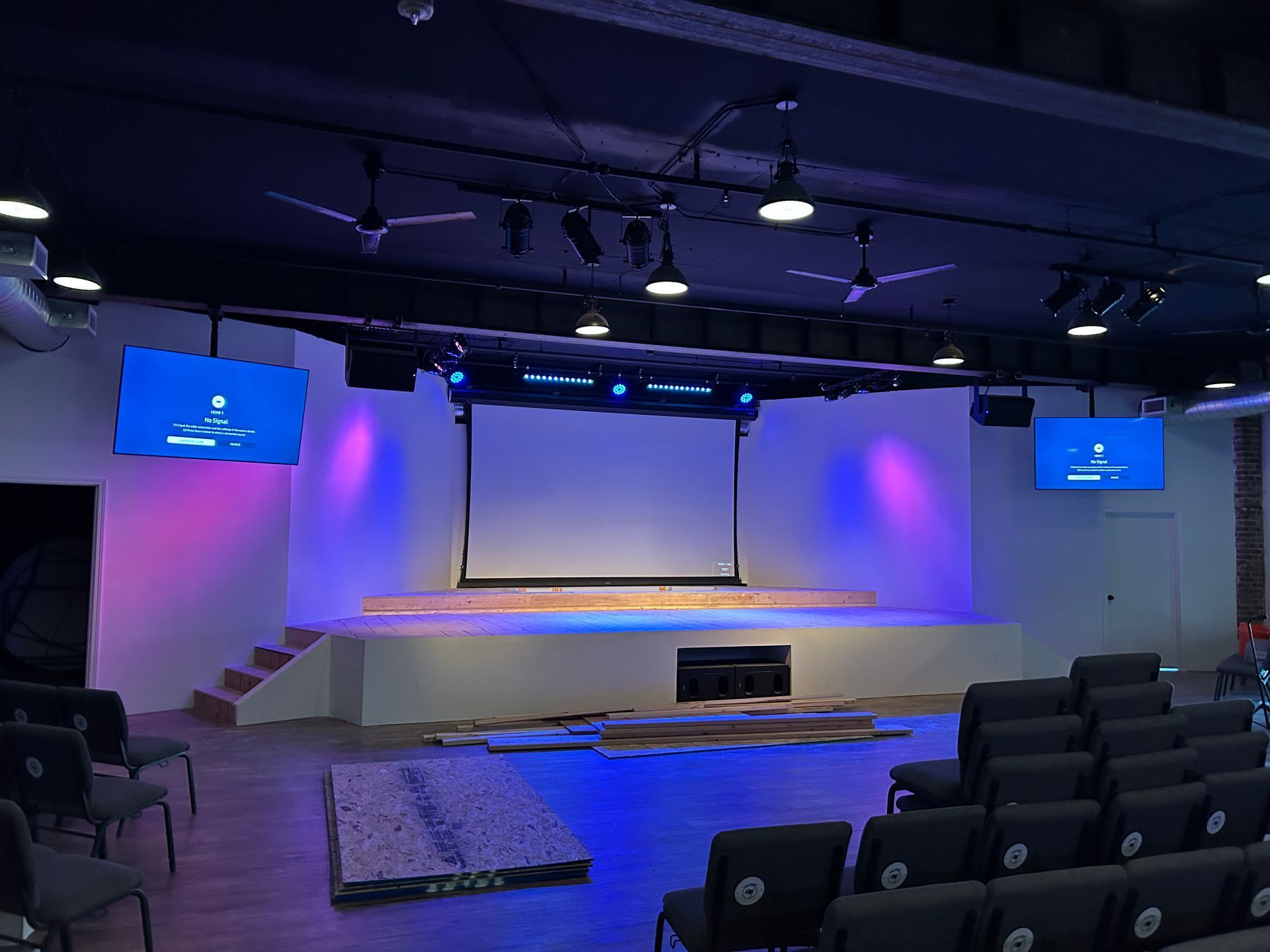 Empty auditorium with purple lighting, stage, projector screen, and rows of black seats