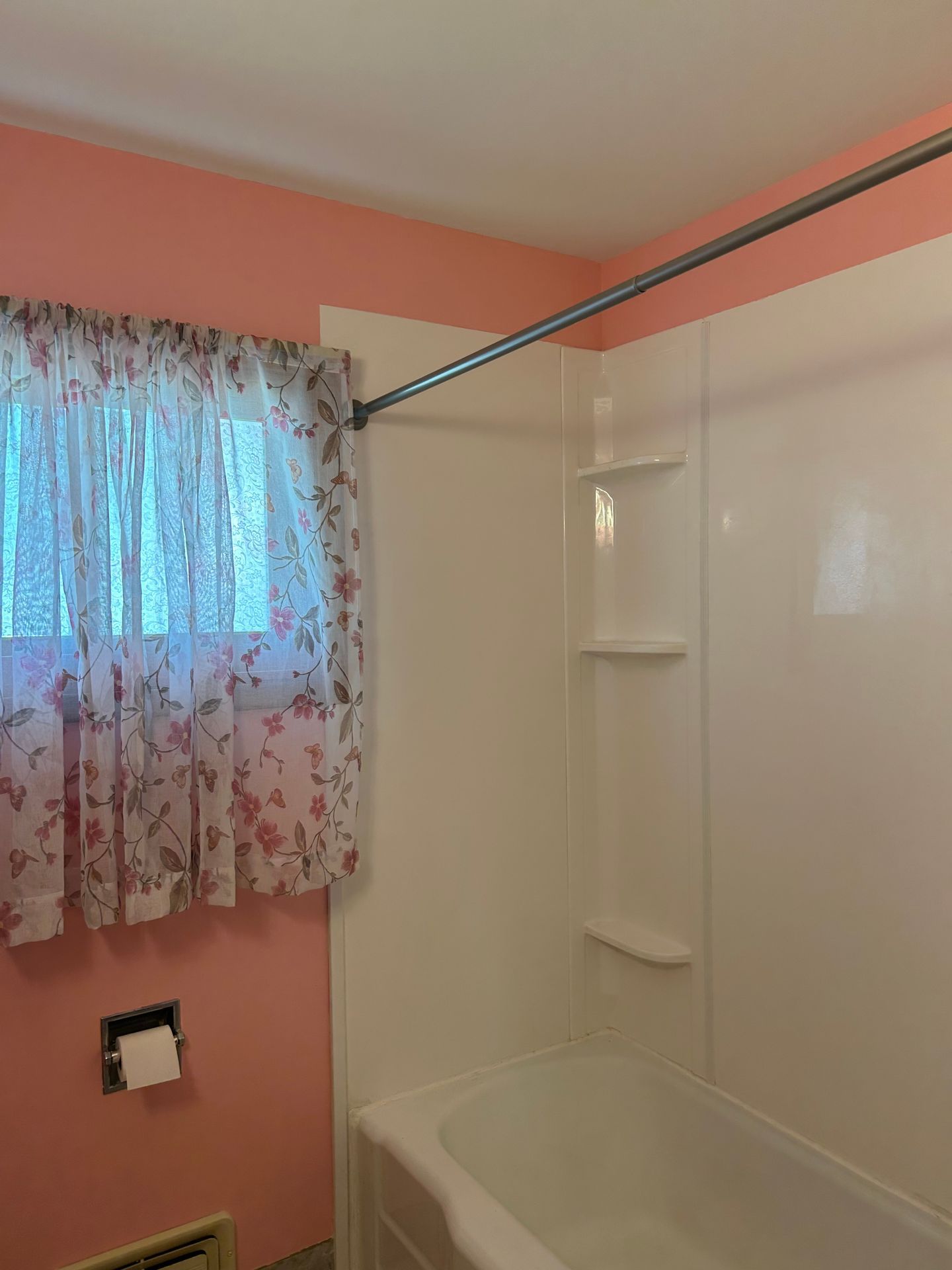 Bathroom with pink walls, white shower surround, bathtub, and a window with floral curtains.
