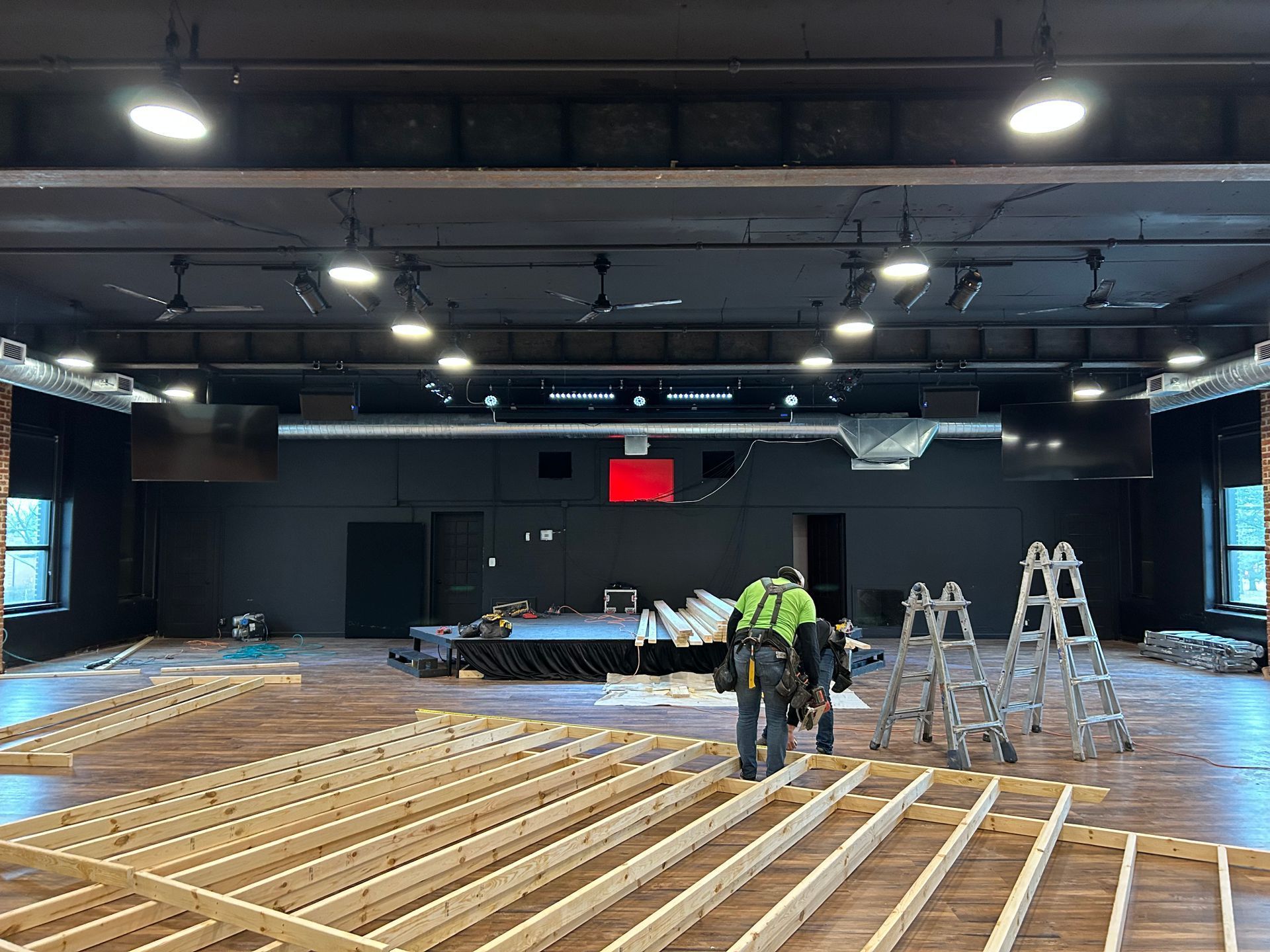 Workers install wooden floorboards in a large indoor hall with ladders and ceiling lights.