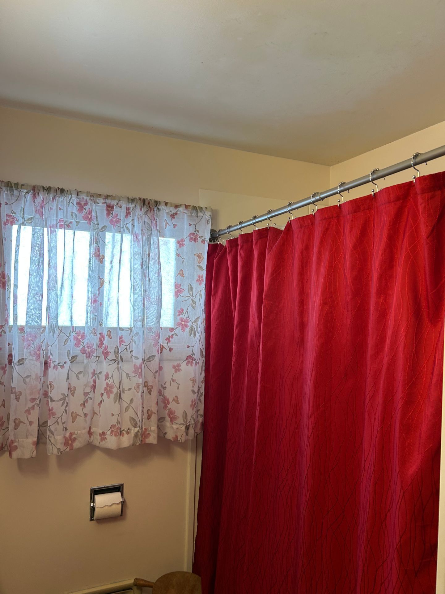 Bathroom corner with beige walls, red shower curtain, and small window with floral curtains