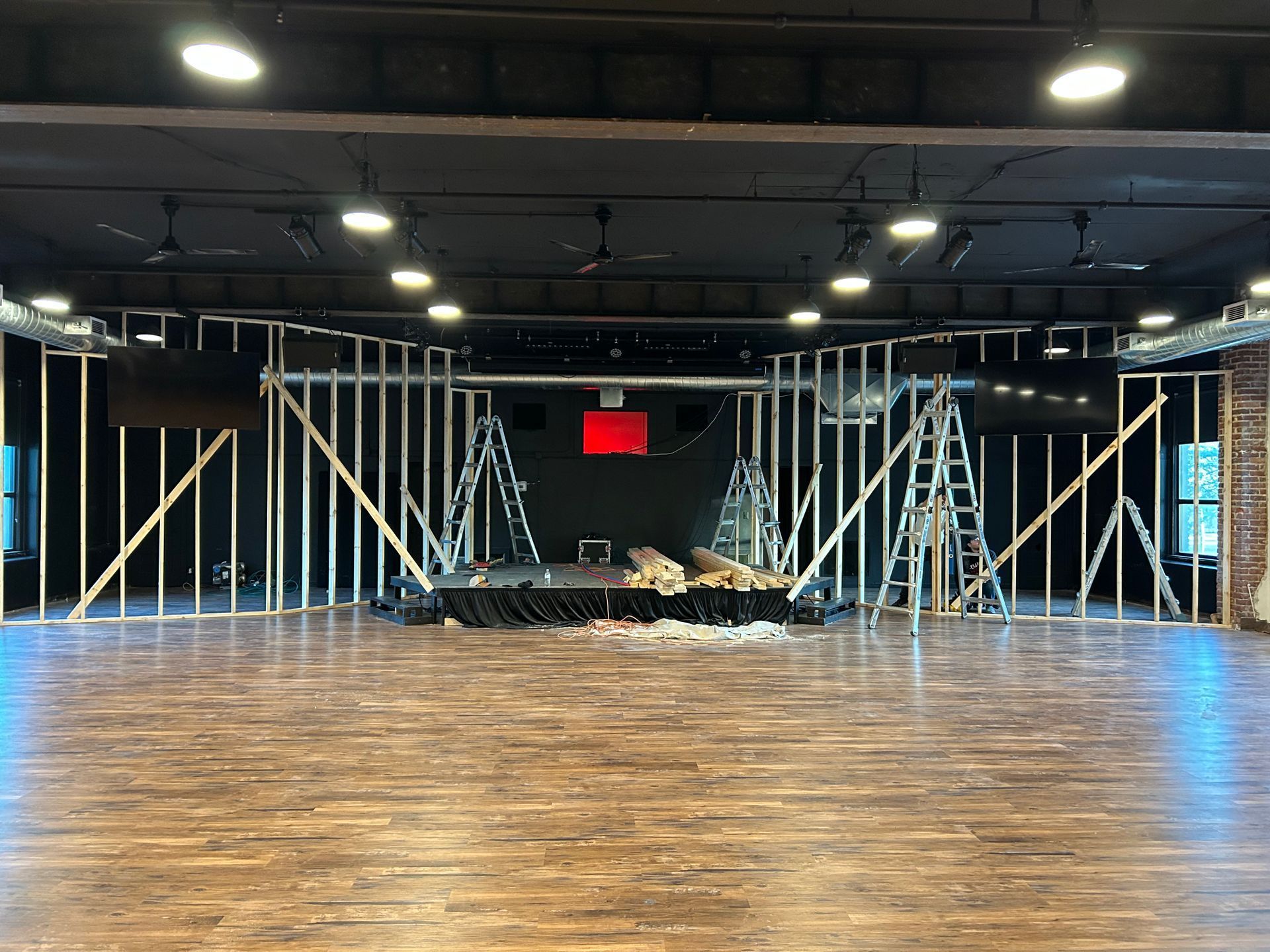 Empty stage with wooden floor, metal lighting truss, and a red EXIT sign at the back