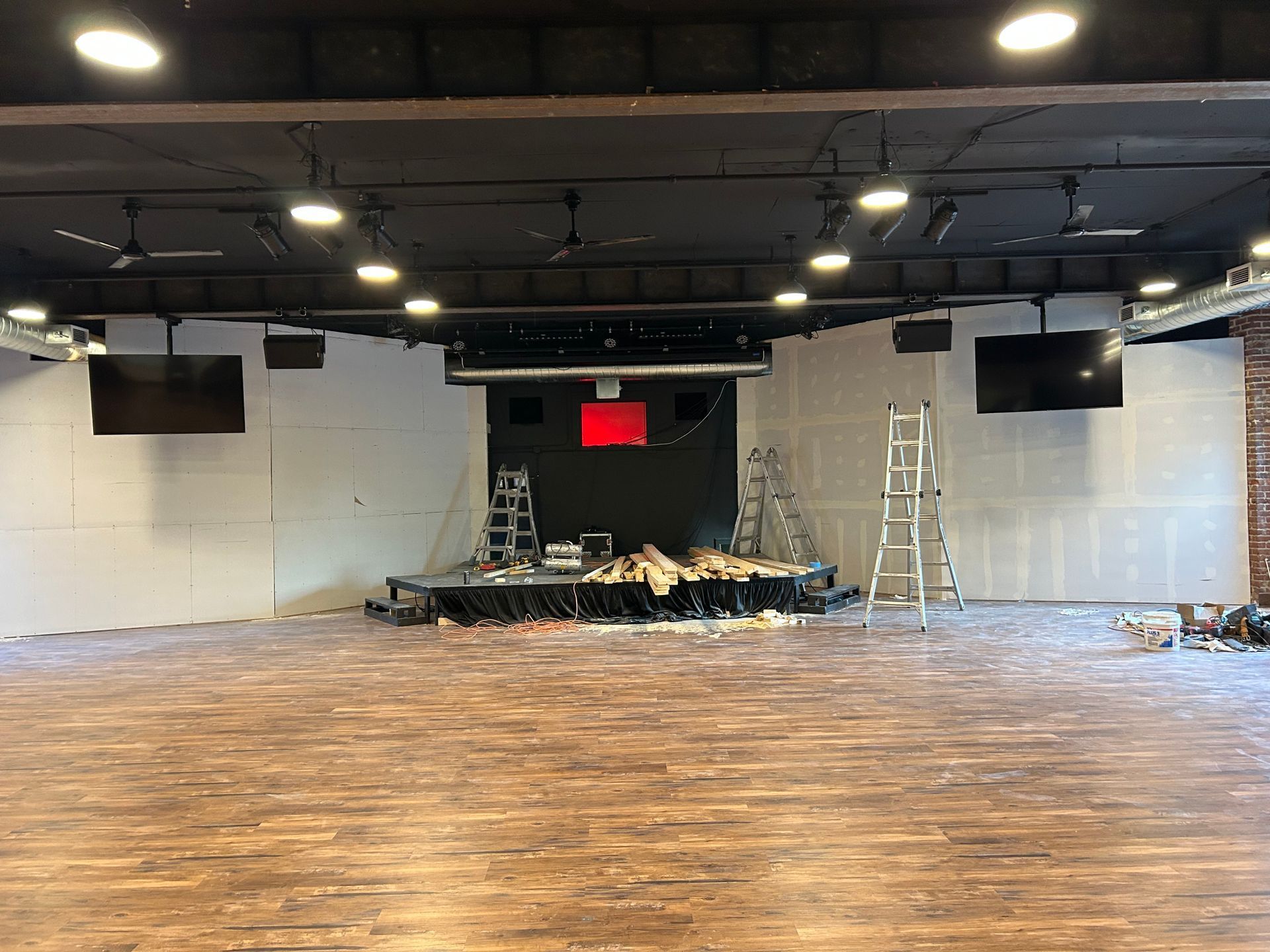 Empty indoor stage with black backdrop, overhead lights, ladders, and a wood floor under renovation