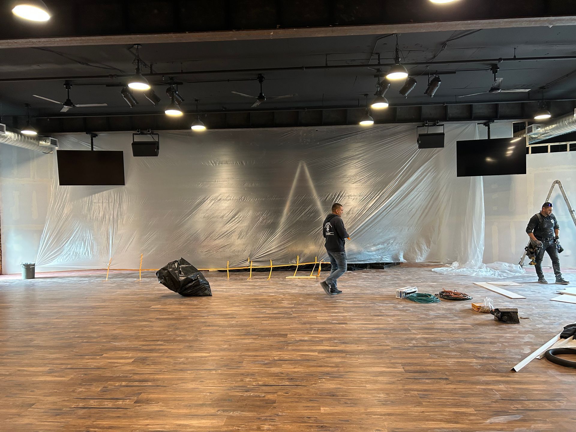 Spacious studio with plastic-covered wall, wooden floor, scattered equipment, and two people standing apart.