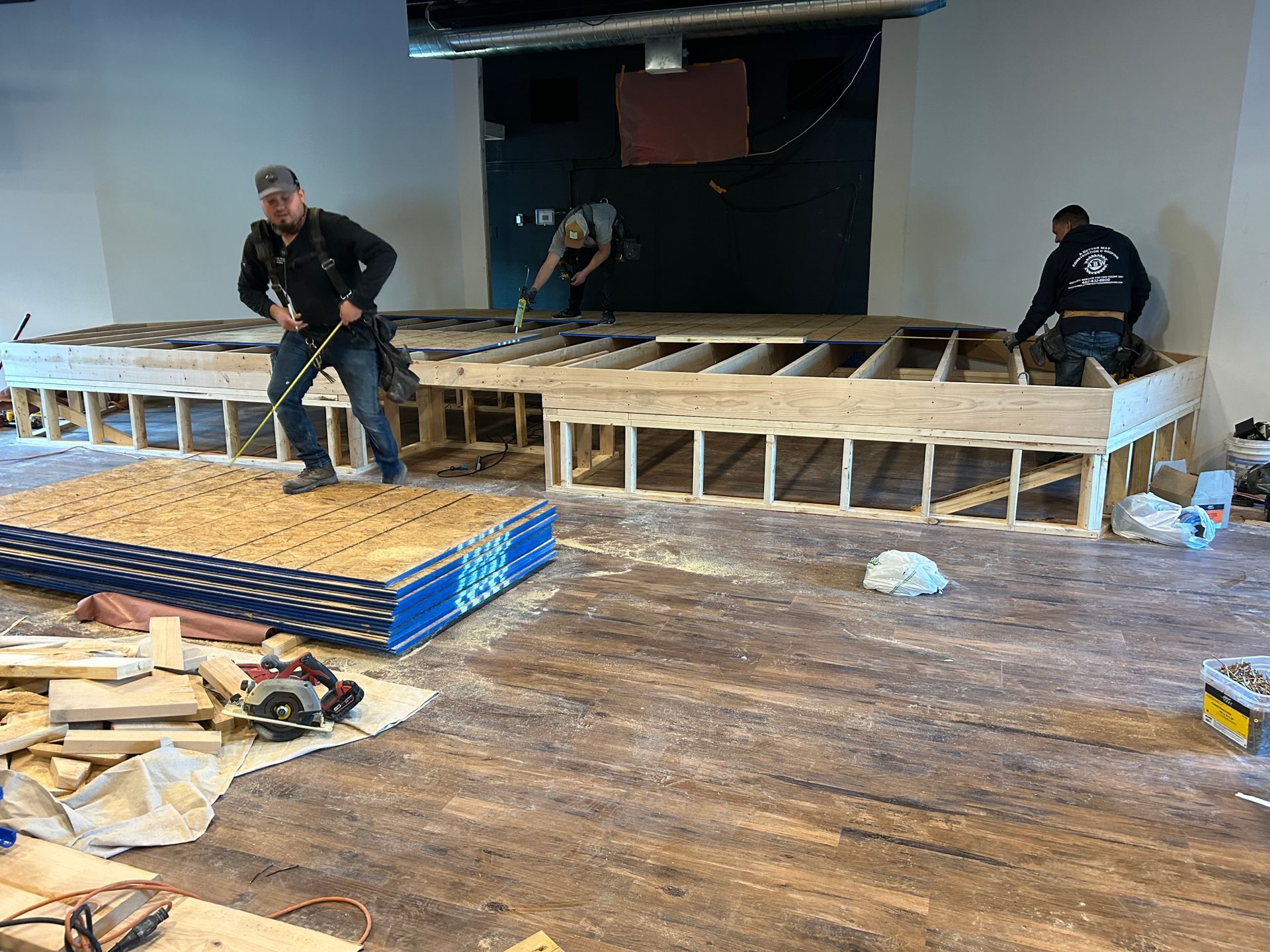 Workers building a wooden stage platform in a large indoor venue, with tools and lumber on the floor.