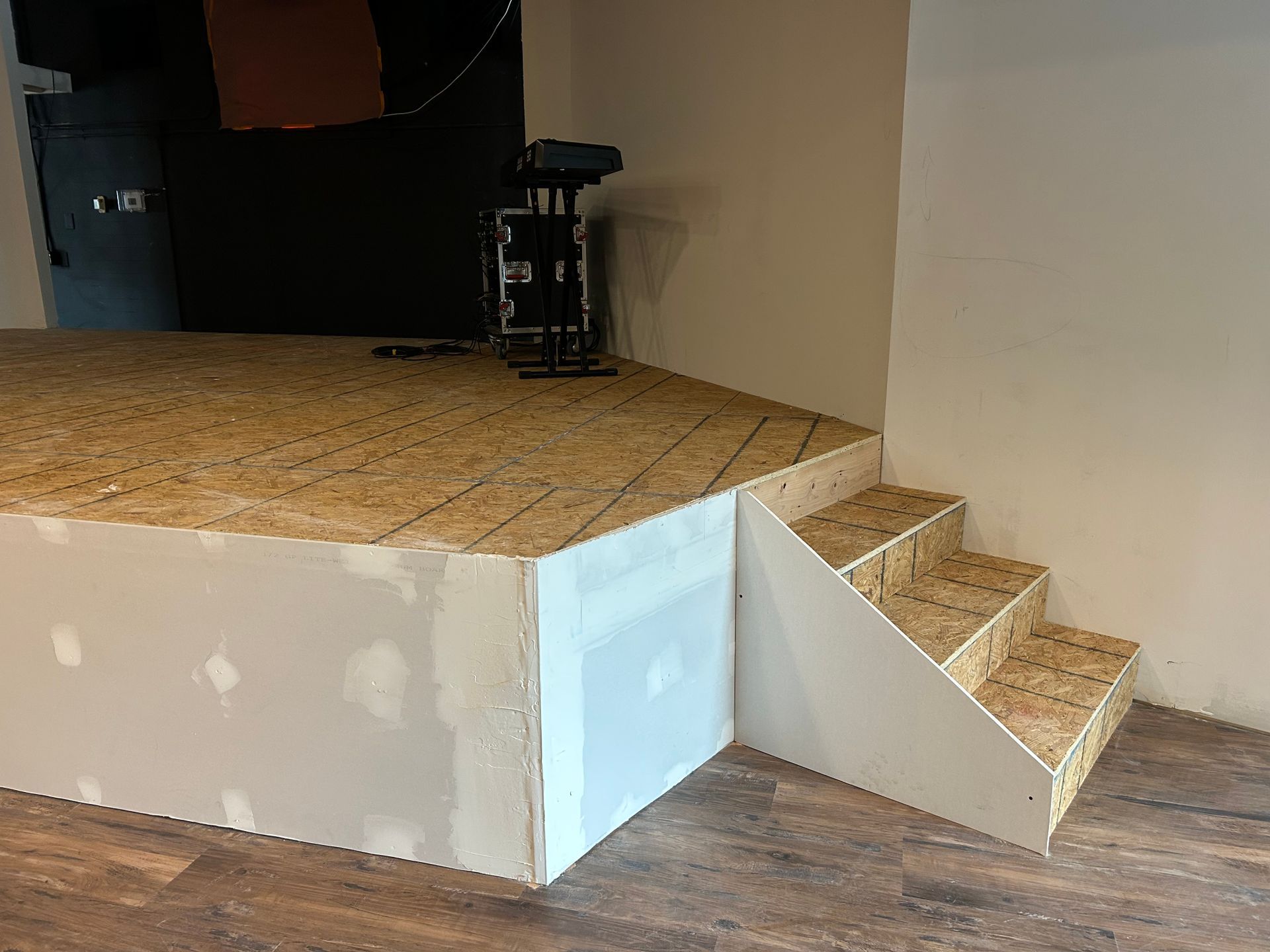 Small stage with plywood floor, white front wall, and short stairs on the right in an unfinished room