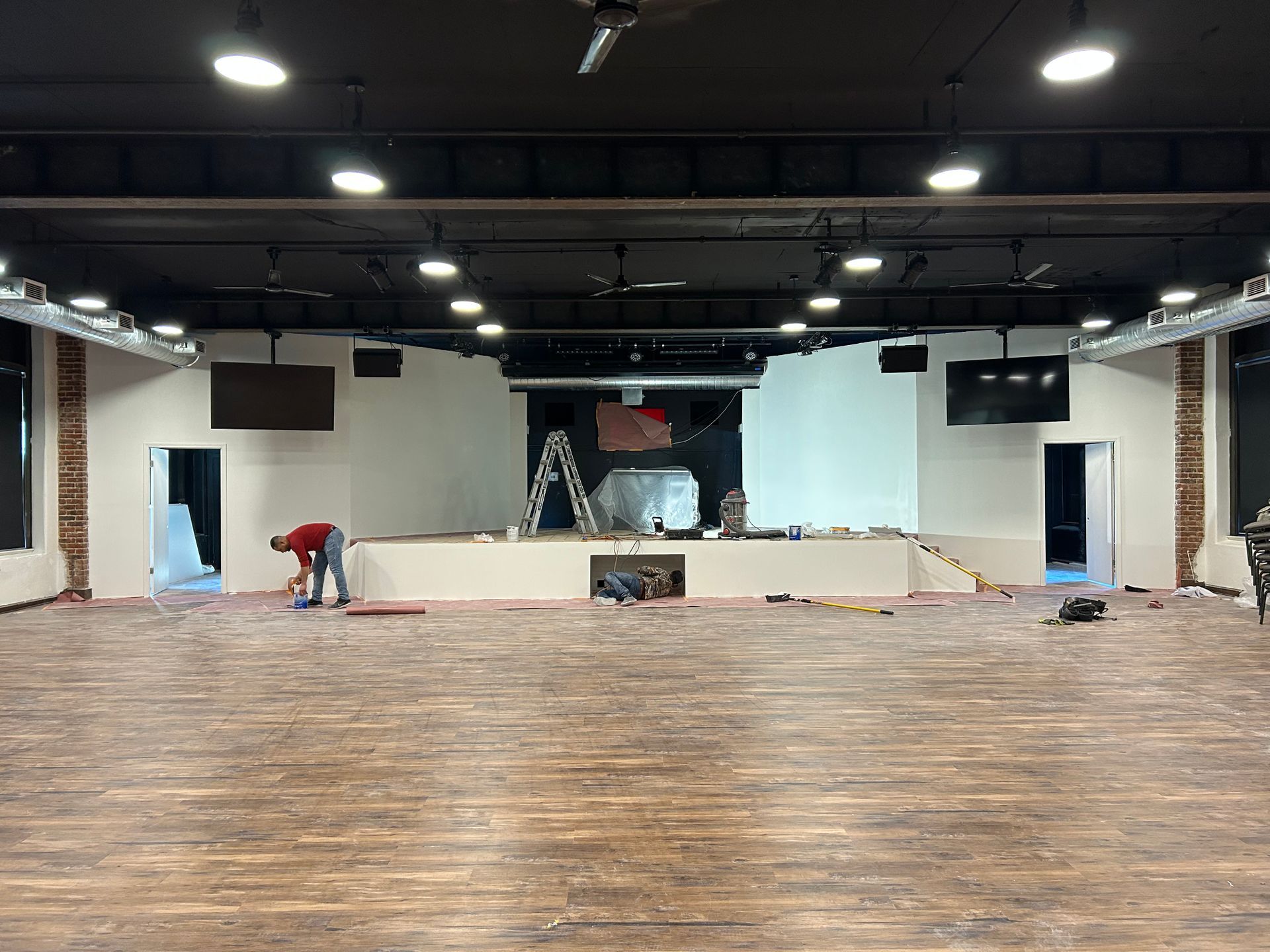 Empty hall with wooden floor, stage, and two workers setting up equipment