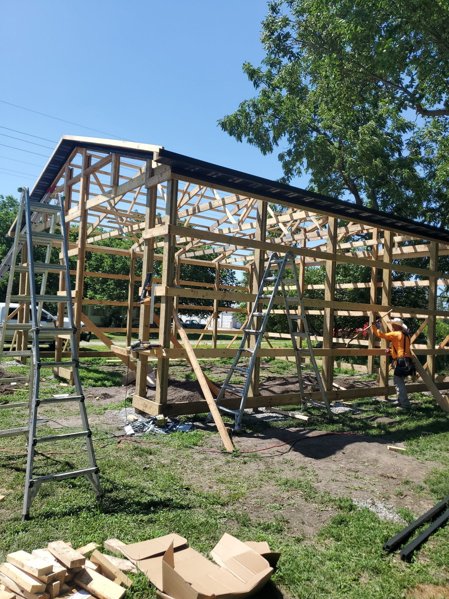 Partially built wooden shed frame outdoors, with a ladder, lumber, and construction materials on grass.