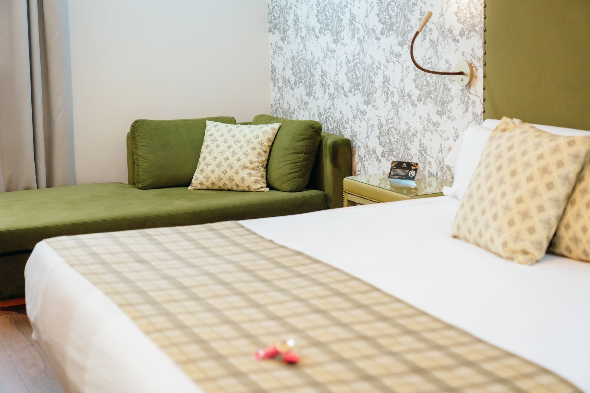 Dormitorio con sofá verde, cama blanca y alfombra a cuadros beige en una habitación luminosa y moderna.