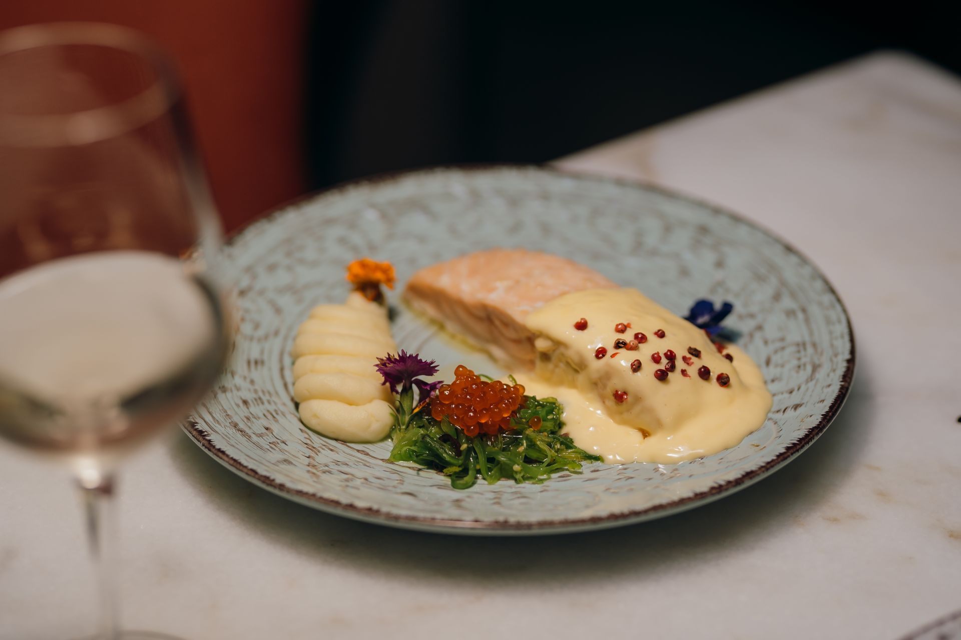 Pescado gourmet con salsa, verduras y caviar en un plato moteado junto a una copa de vino.