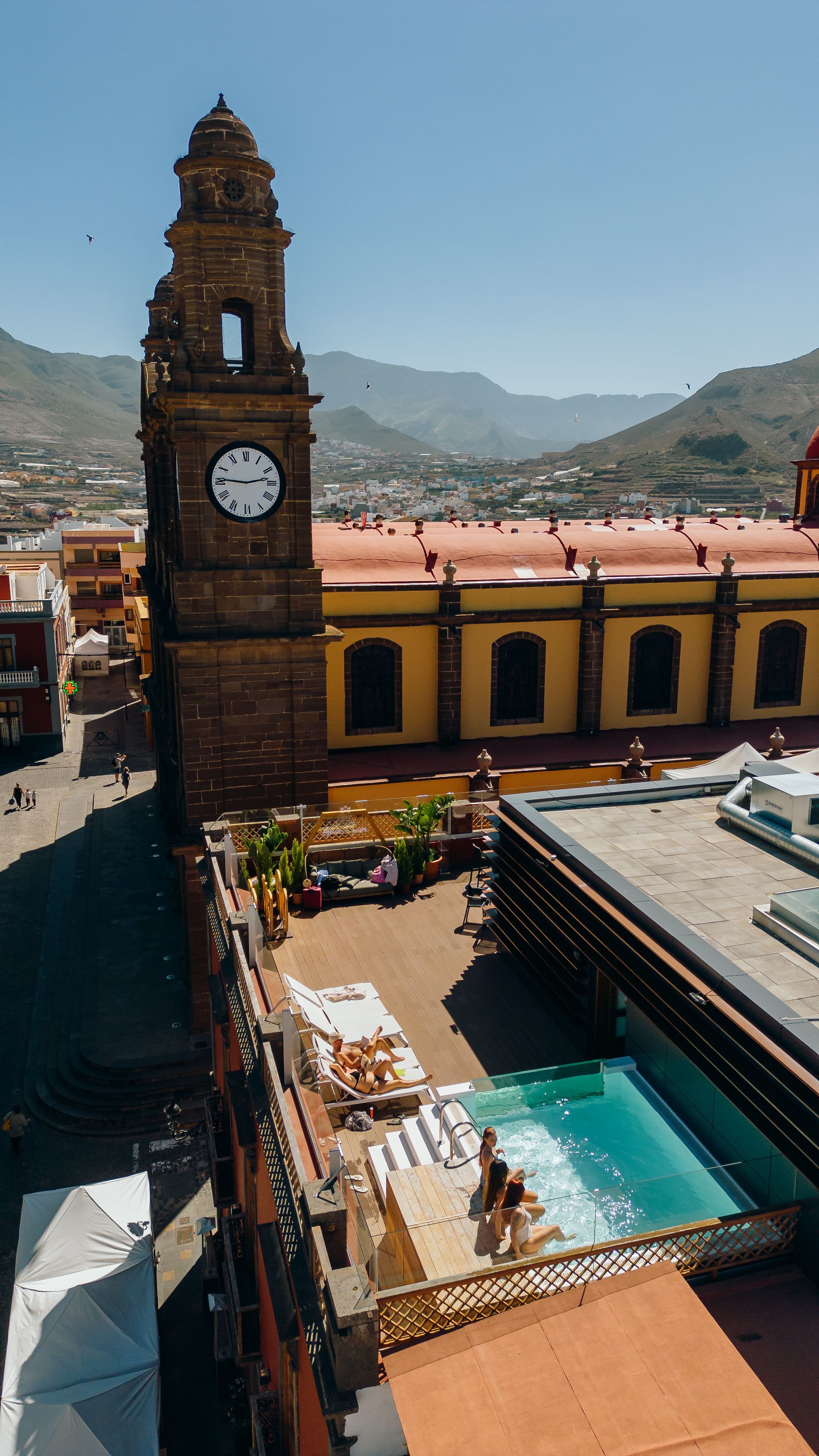 Torre del reloj con vistas a un patio soleado con una piscina turquesa y montañas al fondo.