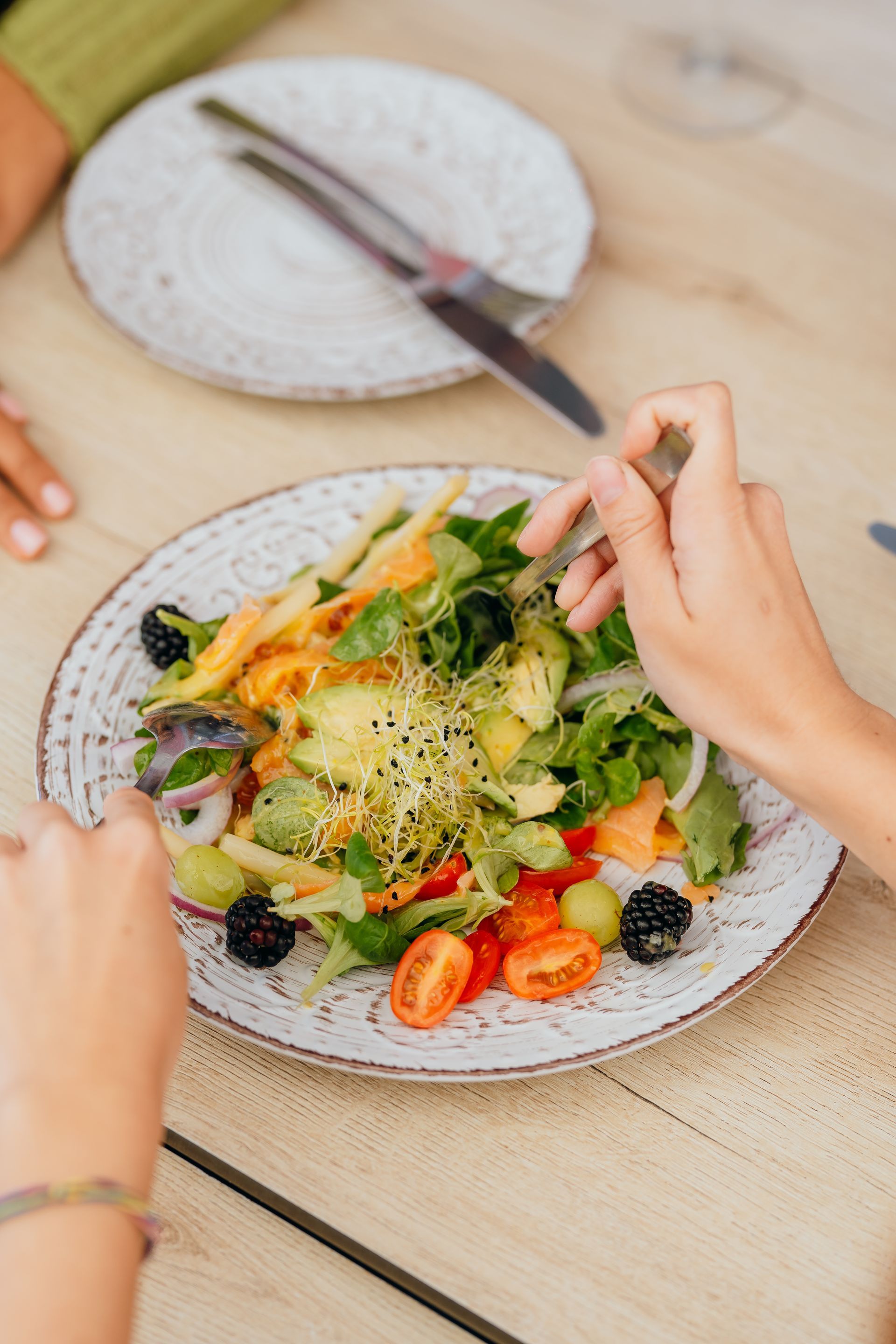 Unas manos mezclan una colorida ensalada con frutos rojos sobre una mesa, con un plato y un cuchillo cerca.